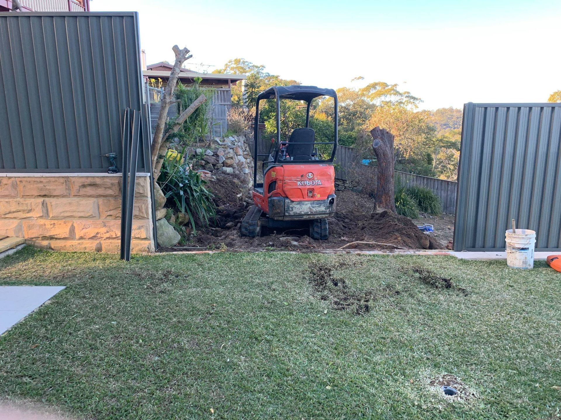 Orange mini excavator working on a dirt pile in a yard, between fences and trees.