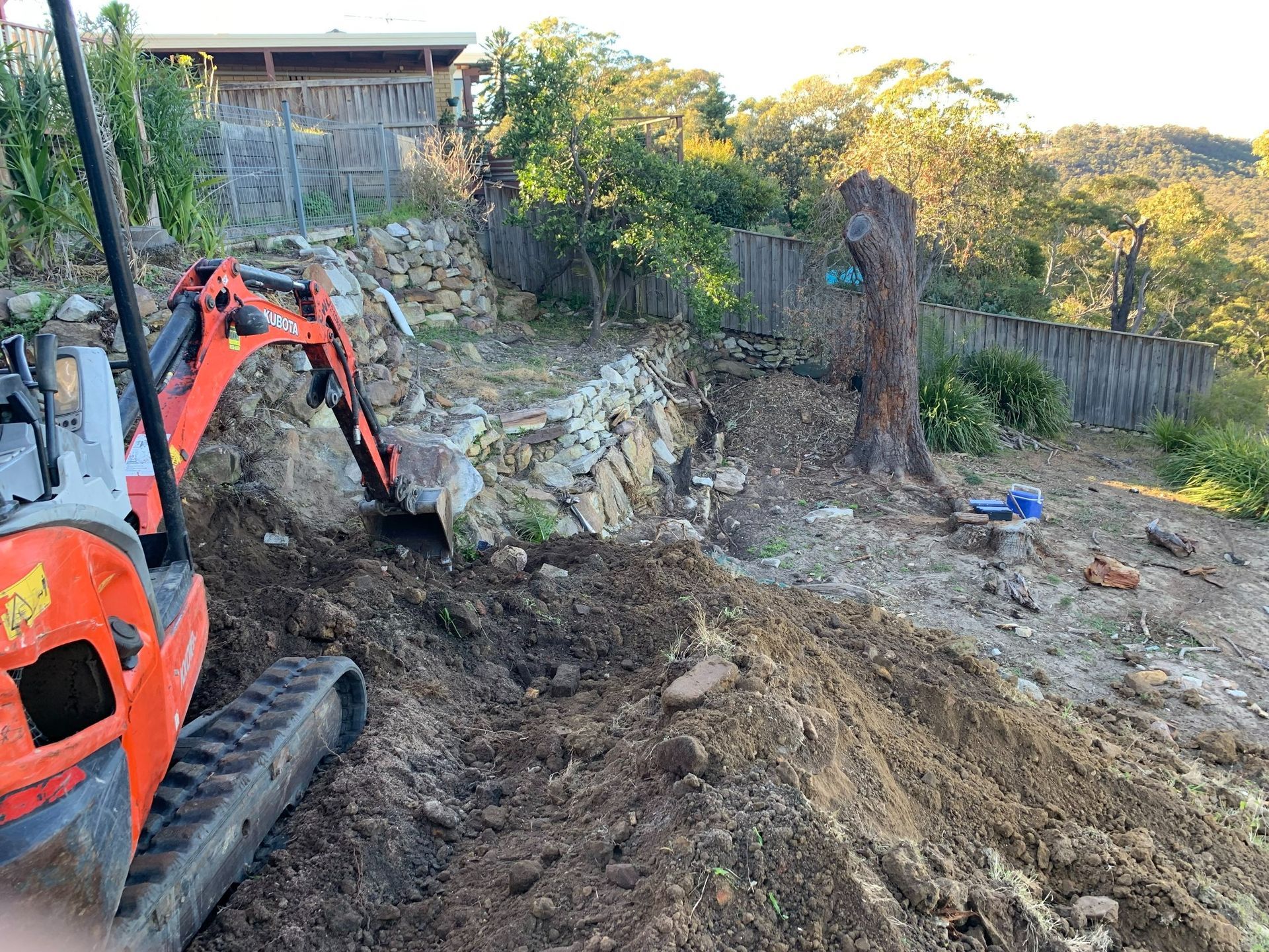 Orange excavator digging into a hillside; a tree stump and fence are visible.
