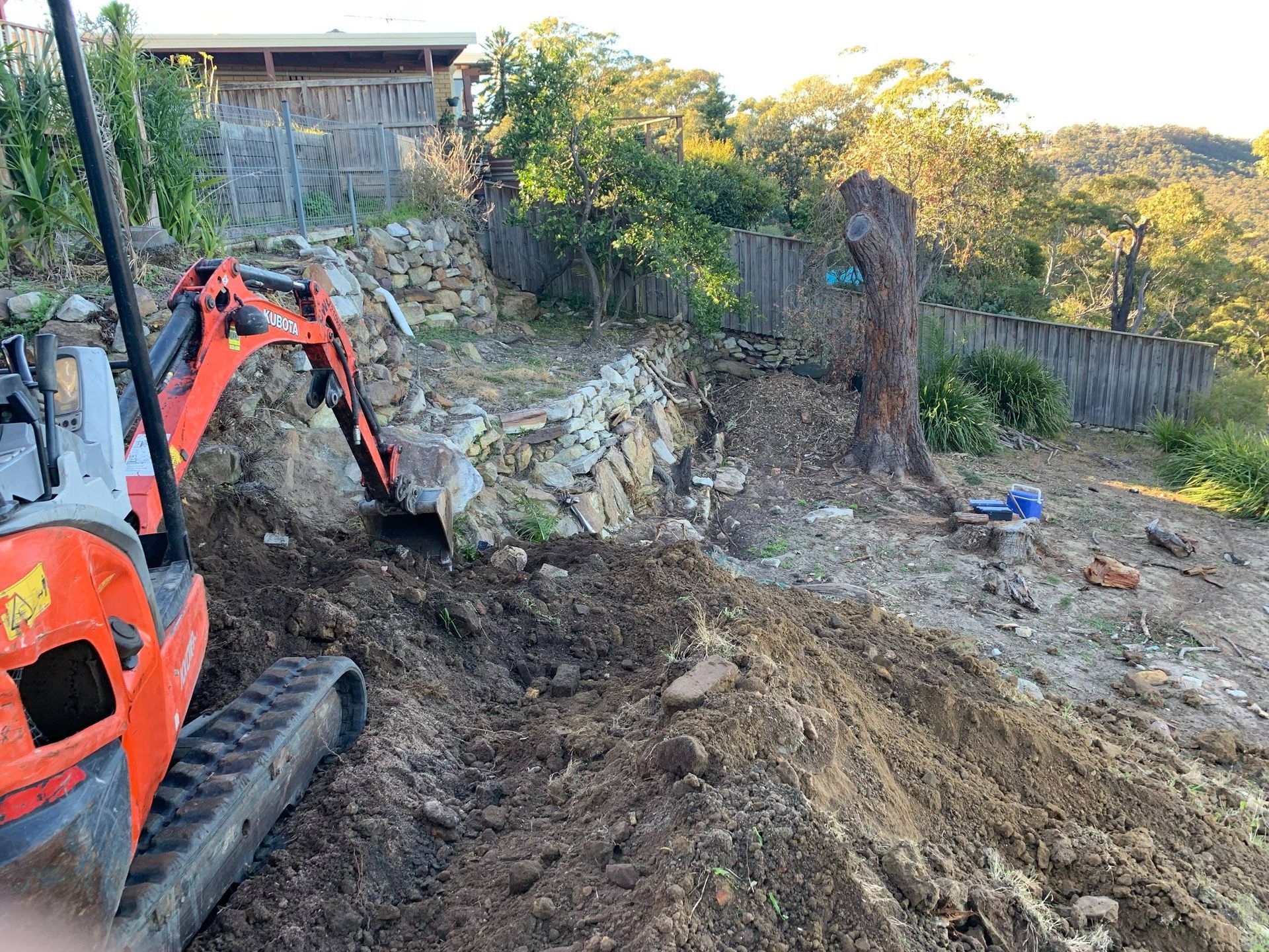 An excavator digs into a hillside with soil and rocks, next to a tree trunk and fence.