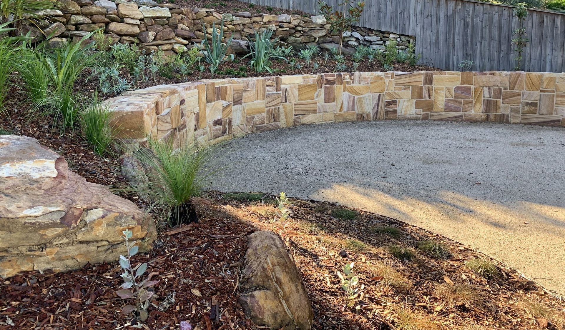 Curved stone wall with gravel surface, landscaping with plants and mulch.