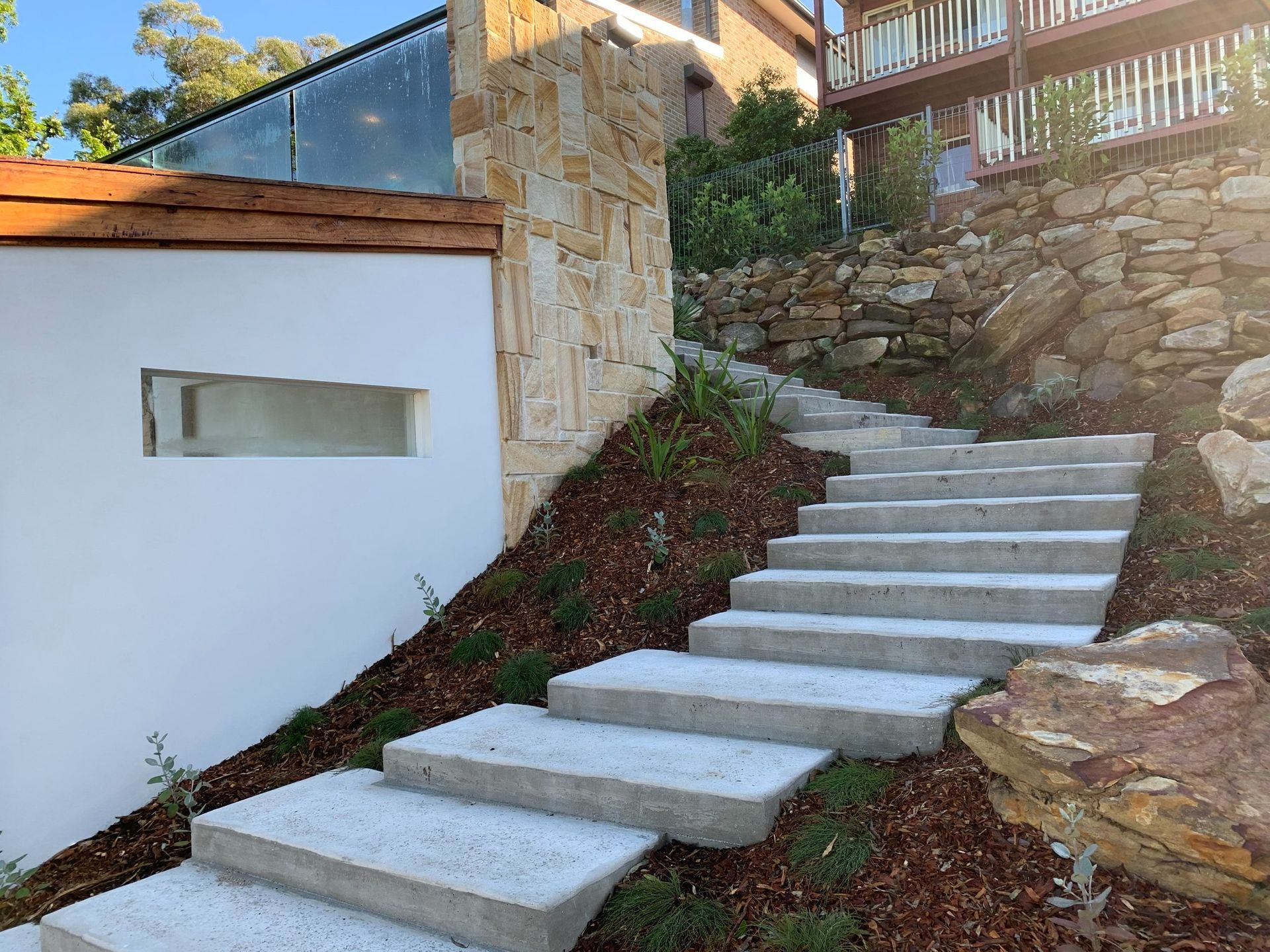 Concrete steps ascend a hillside, flanked by mulch and landscaping, leading toward a building with a stone and white exterior.