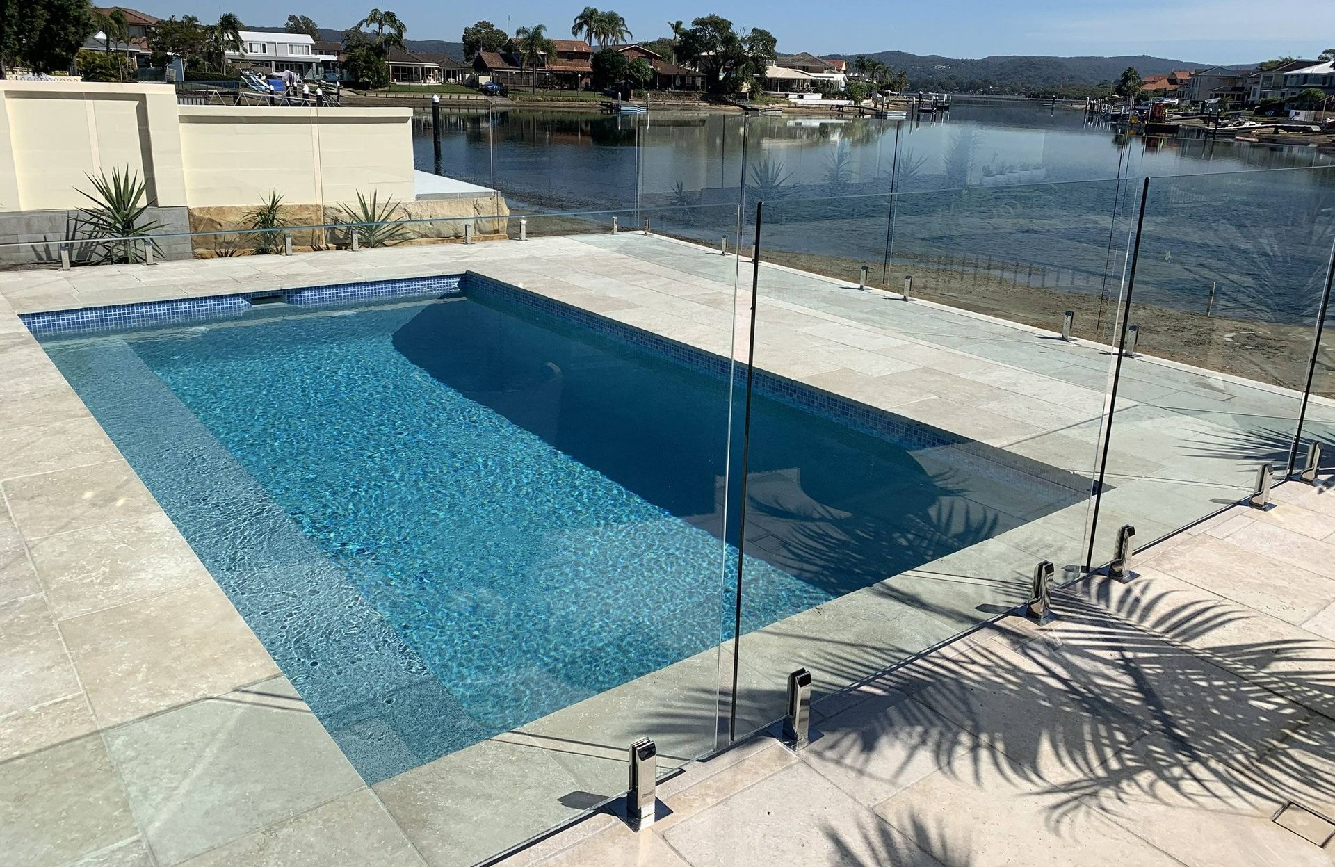 Pool with clear fence overlooking water, buildings in the background. Sunny day.