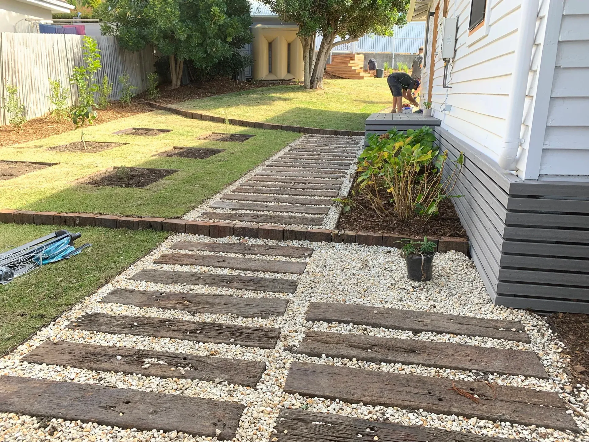 Path of wooden planks and white gravel in a backyard, beside a white house.