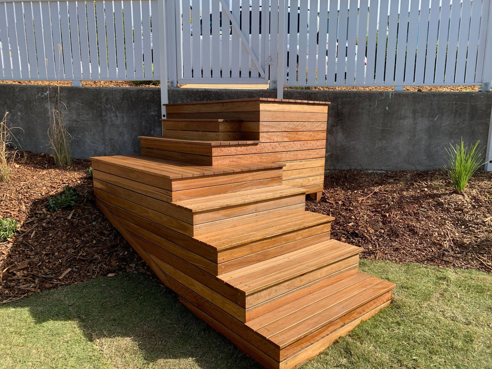 Wooden outdoor steps leading up to a white fence, surrounded by grass and mulch.