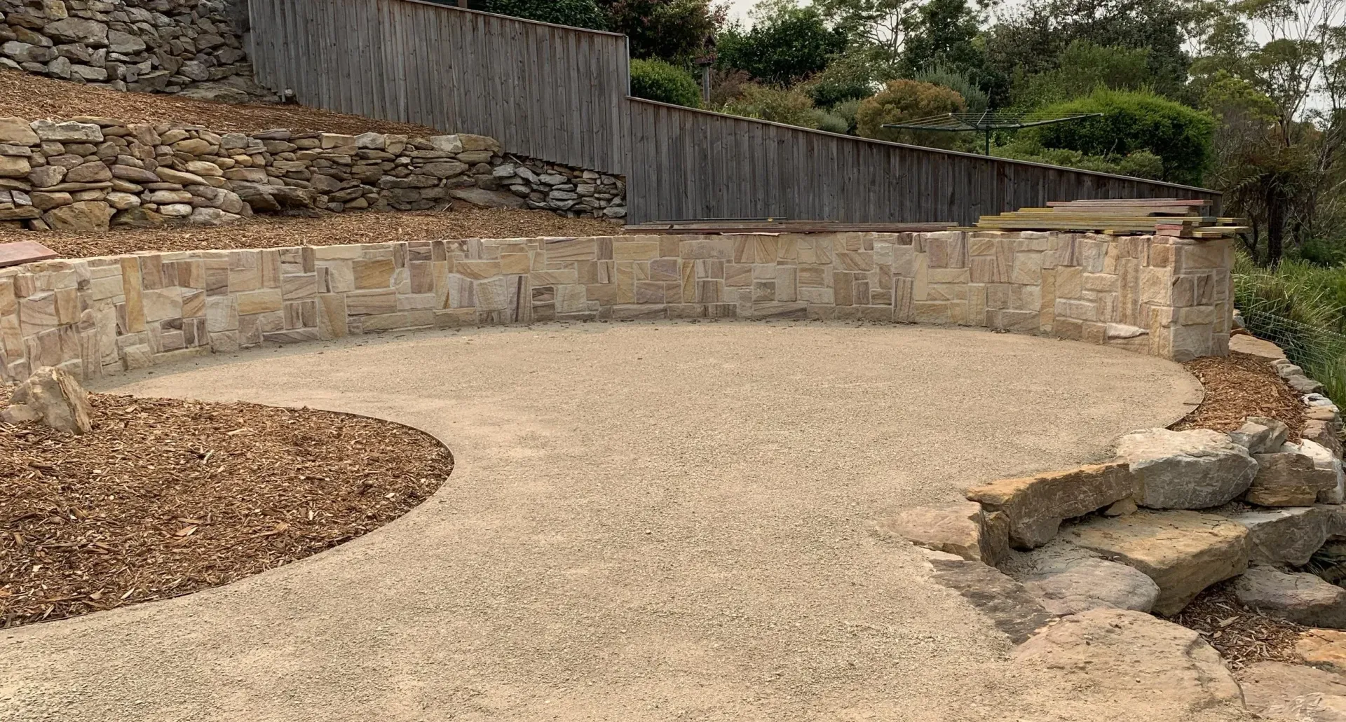 Gravel patio area with stone retaining wall and a wood fence in the background, surrounded by brown mulch.