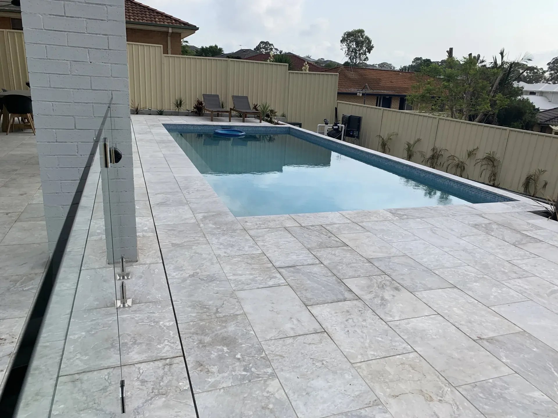 Pool and patio with light-colored stone pavers. Glass railing on the left and a wooden fence in the background.