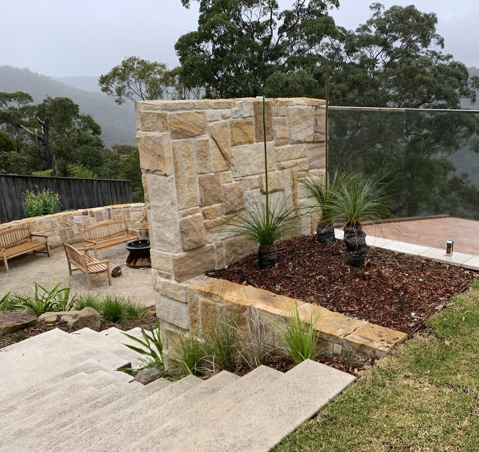 Stone wall with glass panel overlooking a mountainous, forested landscape; stairs in foreground.