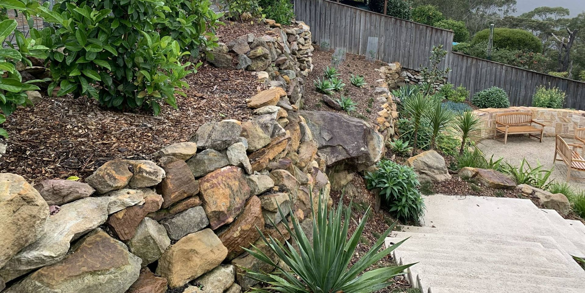 Stone retaining walls with plants and steps leading to a seating area near a wooden fence.