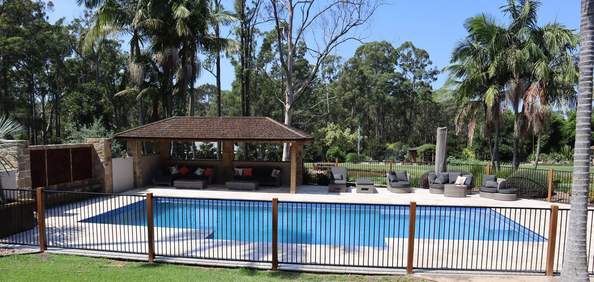 A swimming pool with a gazebo, surrounded by trees and a fence on a sunny day.