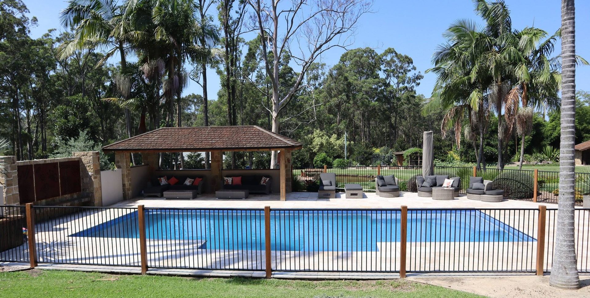 Swimming pool with a thatched-roof gazebo, lounge chairs, and a decorative fence; surrounded by trees.