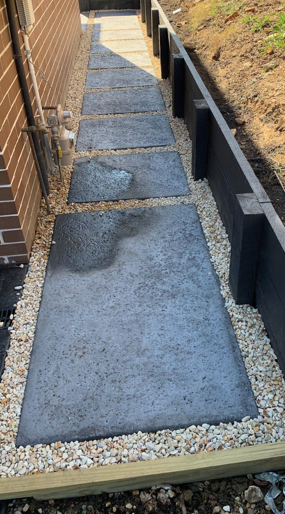Concrete stepping stones in a gravel path bordered by a brick wall and a wooden retaining wall.