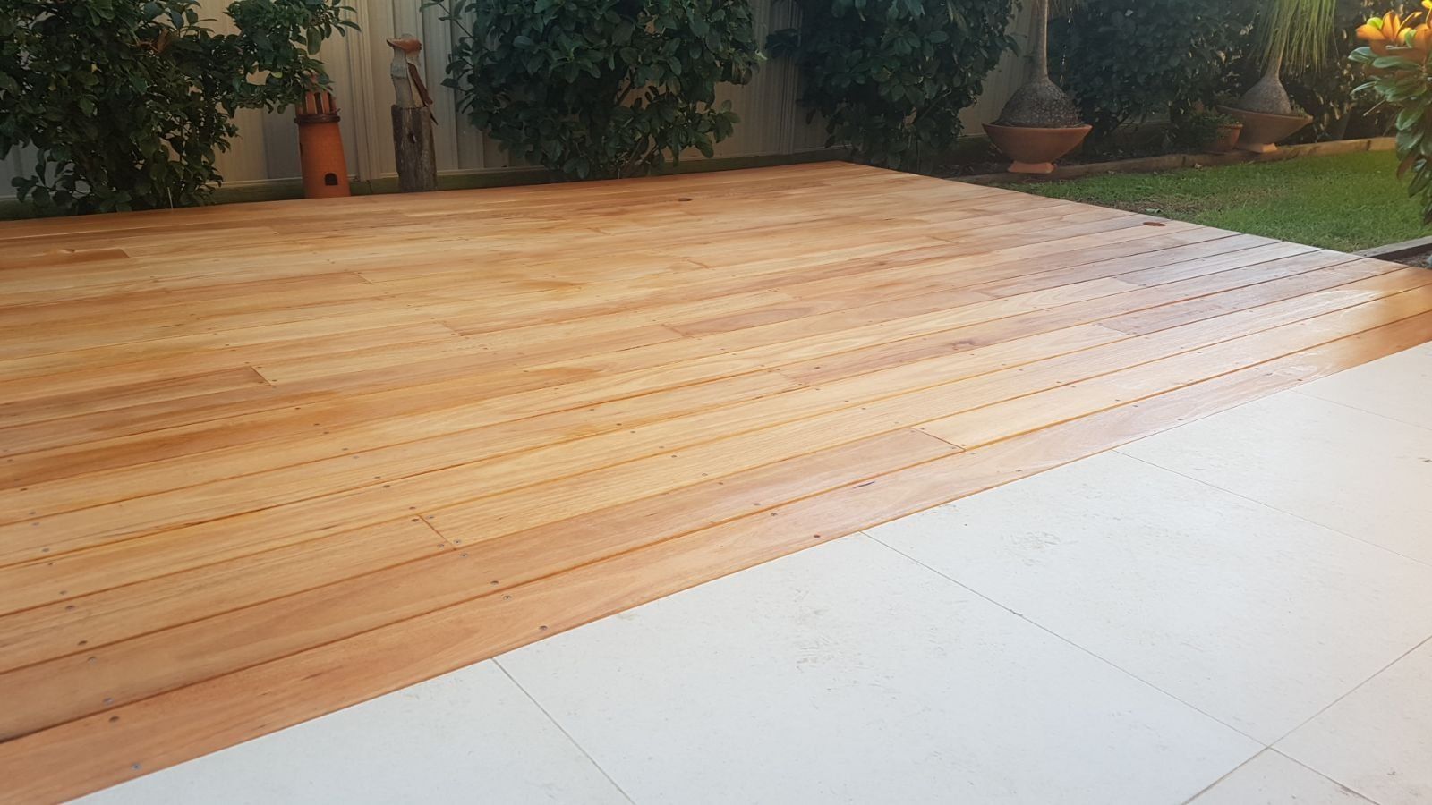 Wooden deck next to white tiled patio, in a yard with green plants and lawn.