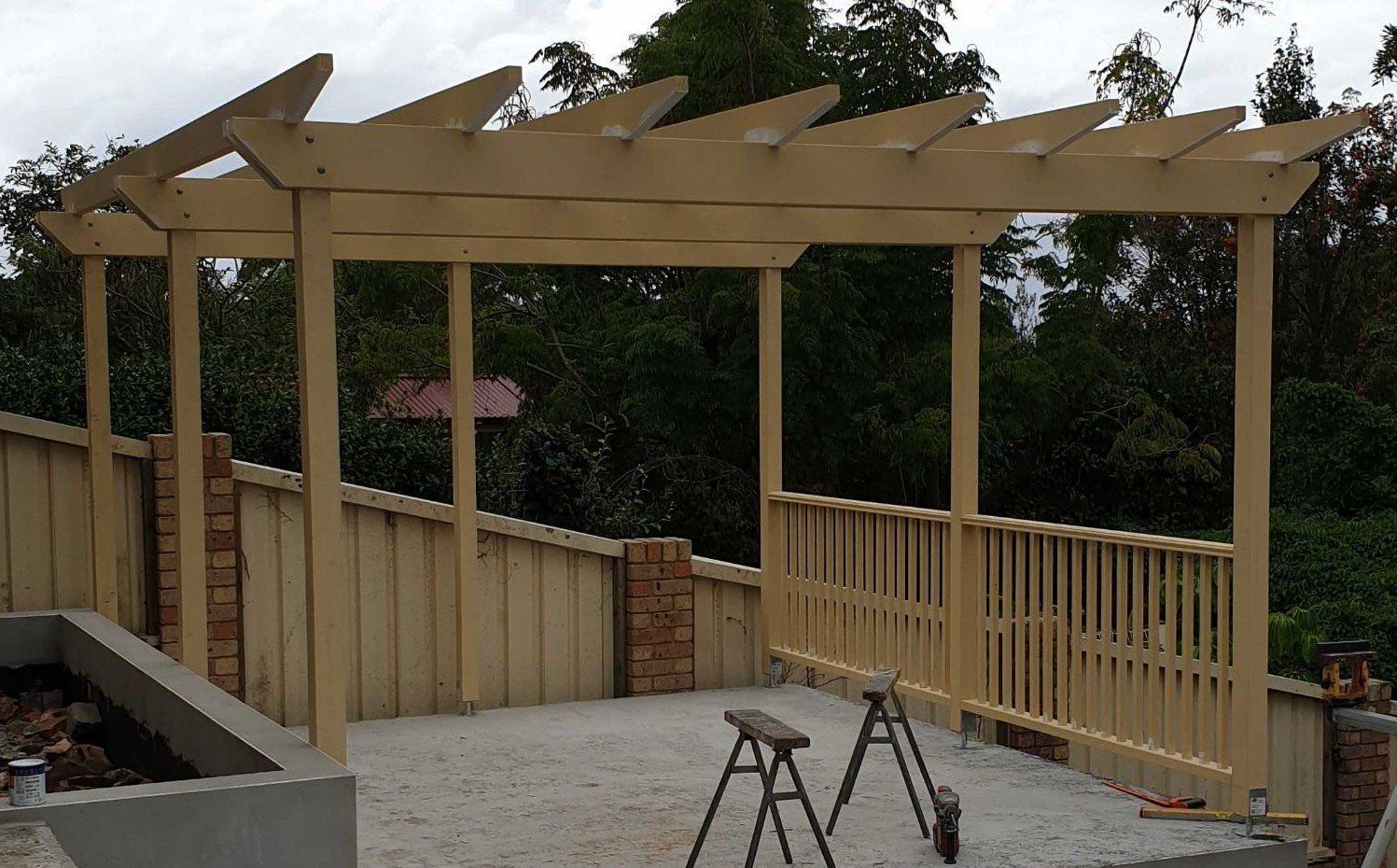 Wooden pergola over a patio, supported by posts and a fence, set against trees.