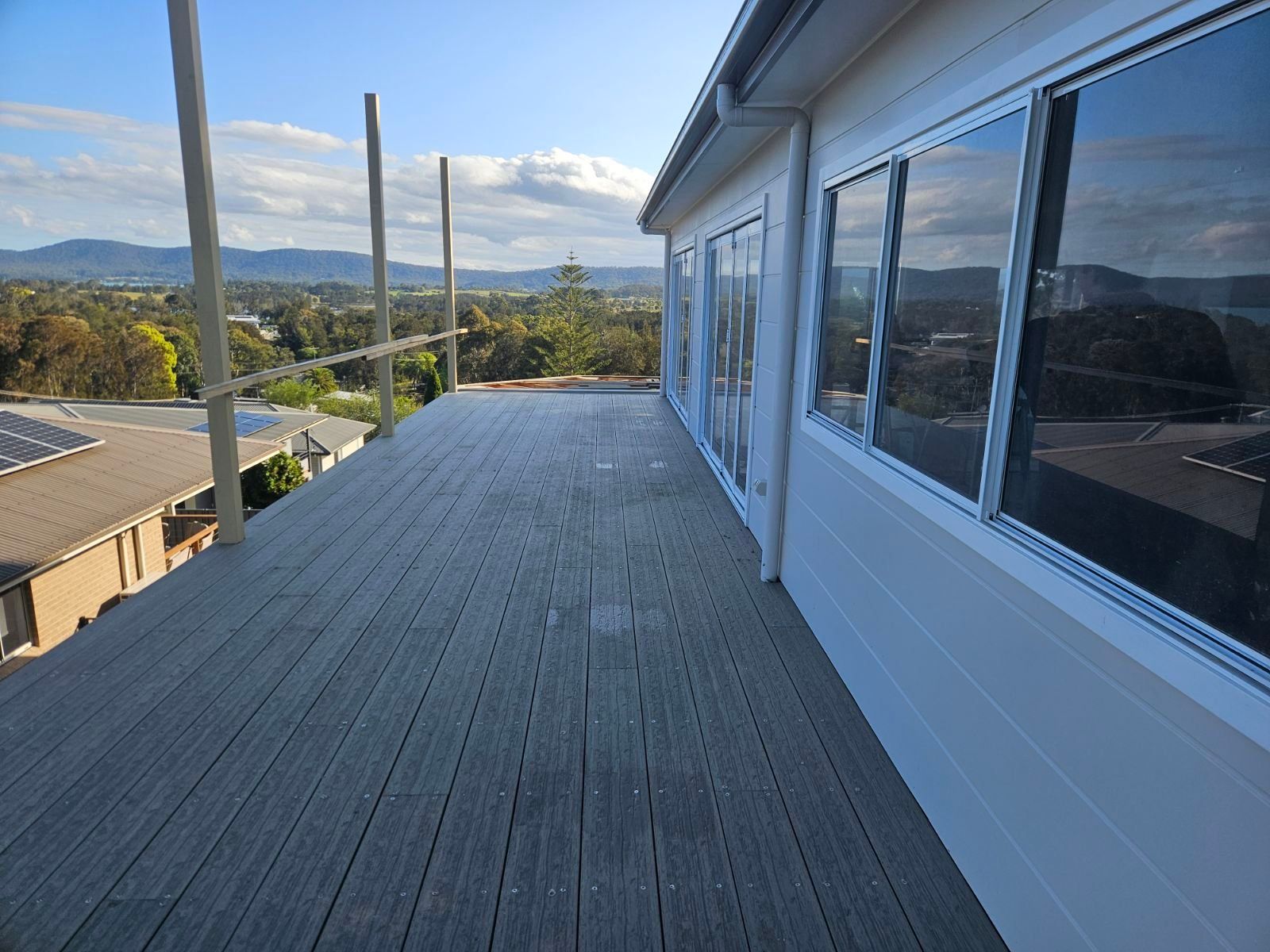 Deck with weathered gray boards, next to a white house with windows. Scaffolding visible. Green trees and hills in the background.