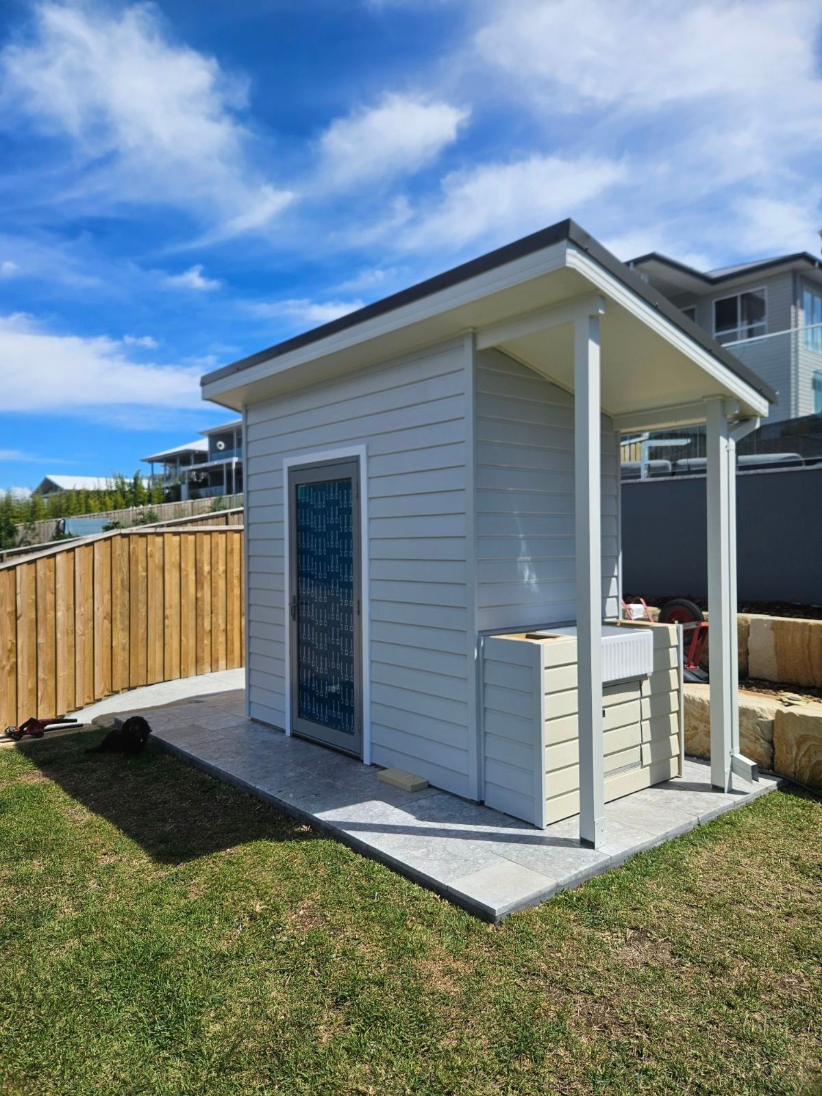 Small outdoor shed with light gray siding and a gray tile patio on a sunny day.