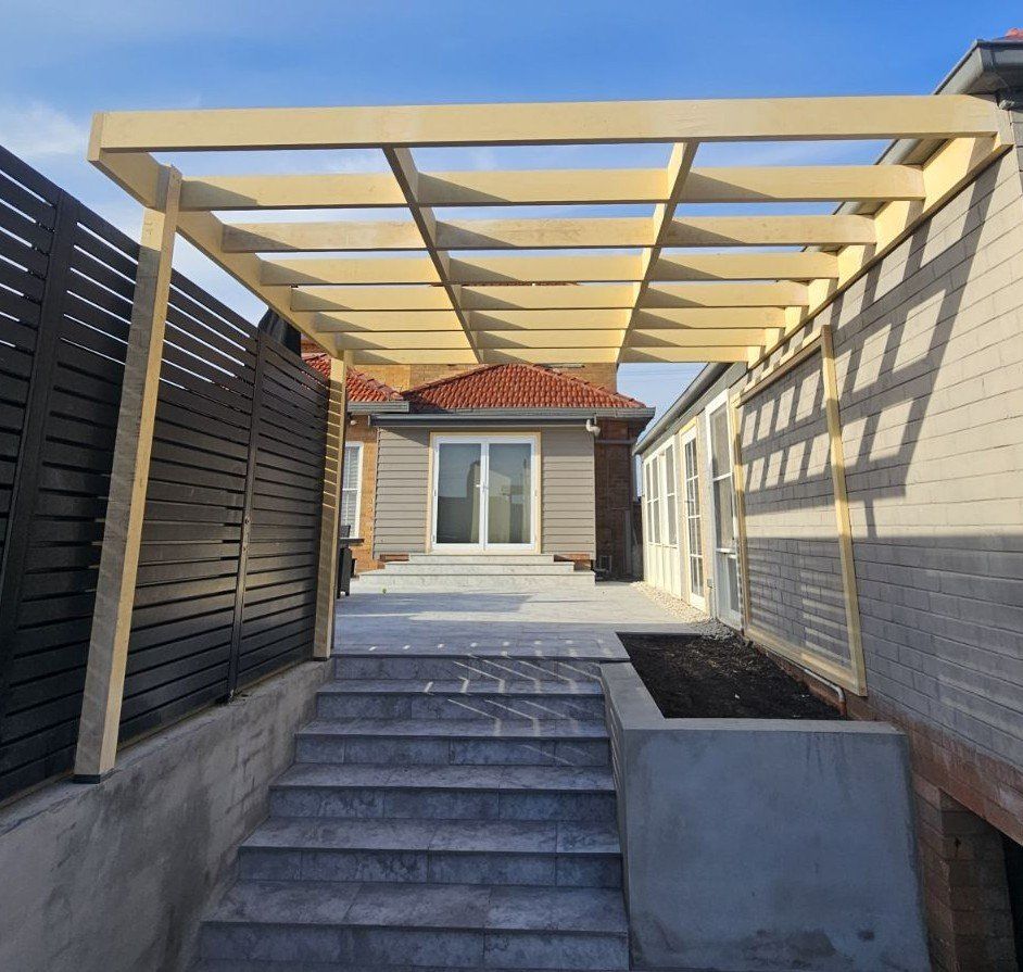 Wooden pergola over concrete steps leading to a house; dark fence on the left.