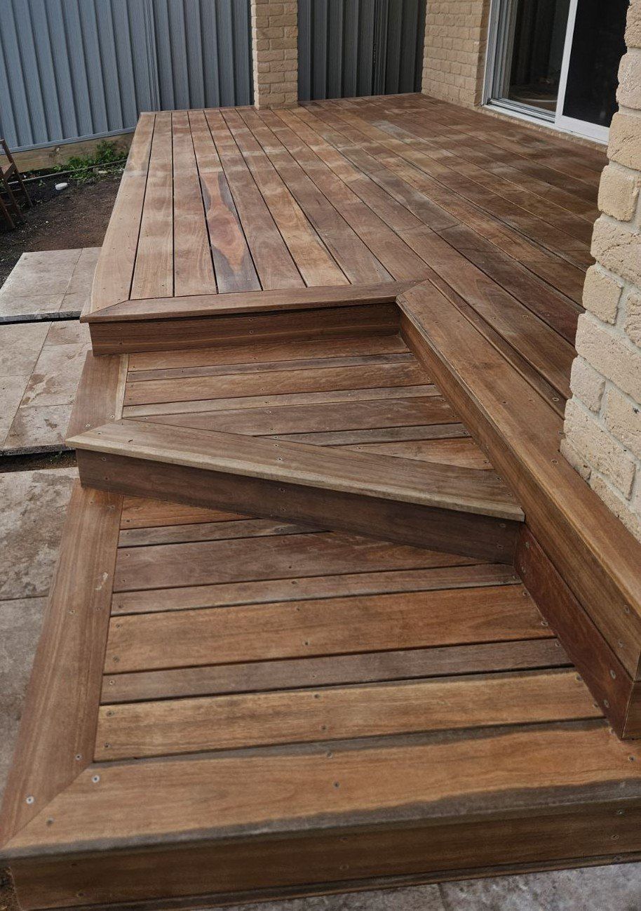 Wooden deck with steps leading to a raised platform against a brick wall.