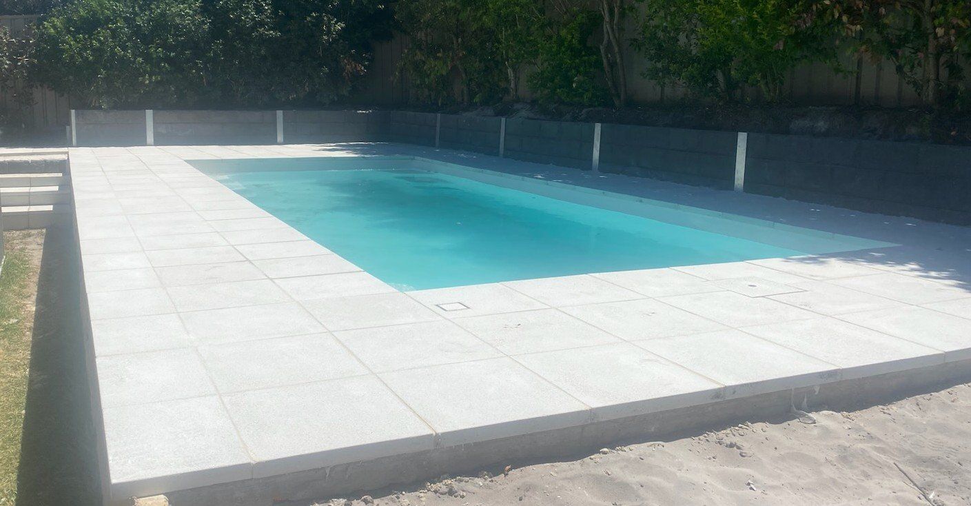 Rectangular swimming pool with gray tiled deck and dark wall, surrounded by green grass and trees.