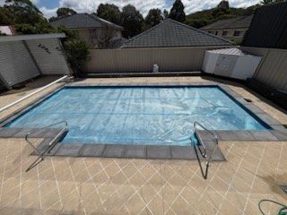Rectangular pool covered with blue material, surrounded by beige tiles and fencing.