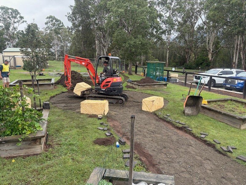 An excavator digs in a garden bed. Gravel path is being laid; blocks of stone present.