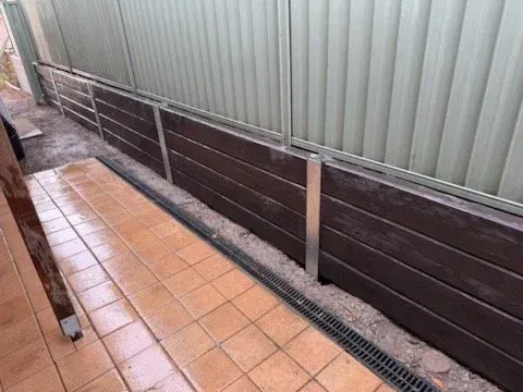 A brown composite retaining wall with metal posts and a corrugated green fence.  A paved area with a drain is in front.
