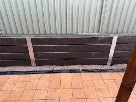 Brown wooden retaining wall with metal supports, against a corrugated metal fence, over a tiled floor with a drainage grate.