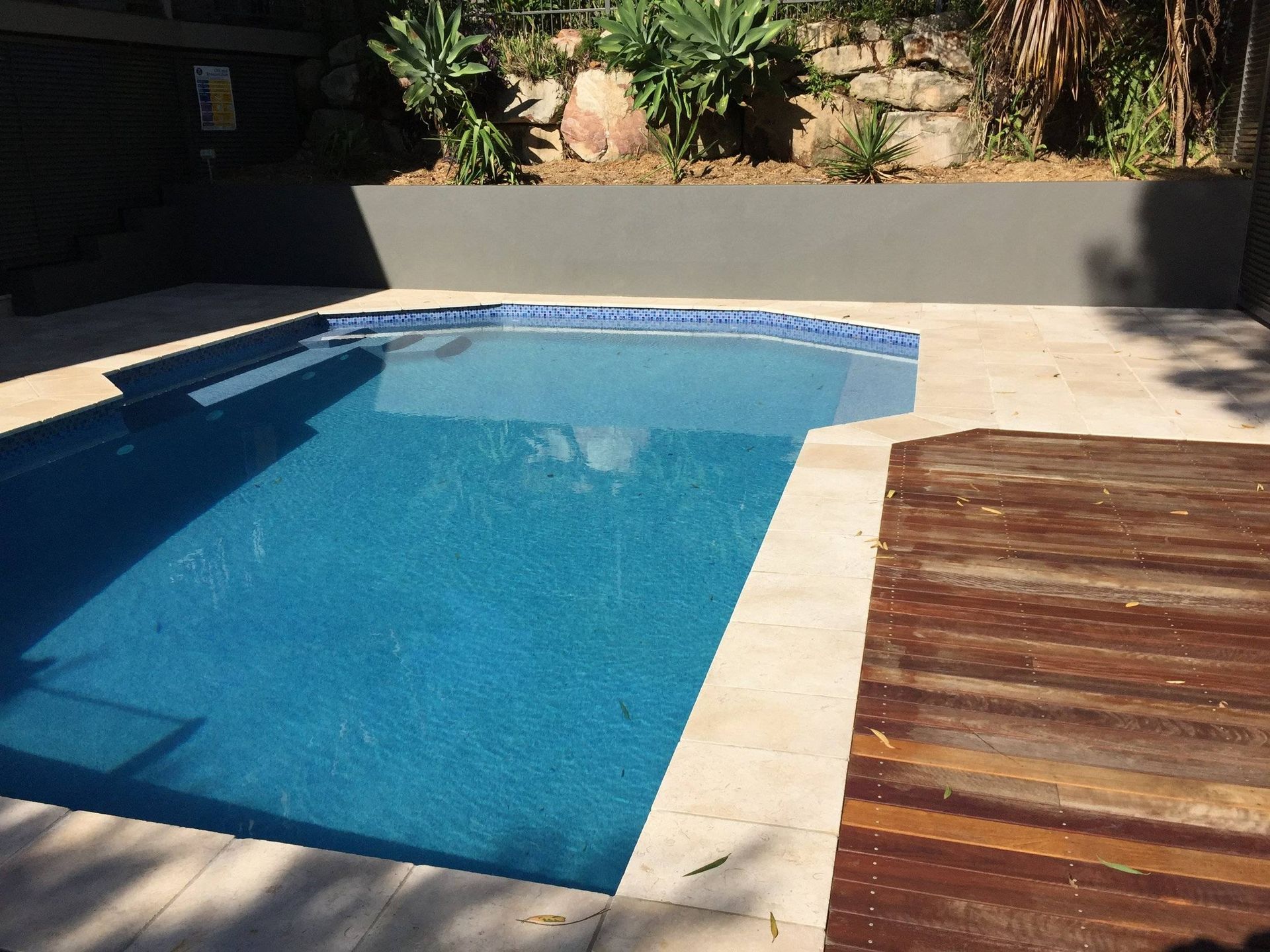 Swimming pool with blue water and stone surround, next to wooden deck and planted garden.