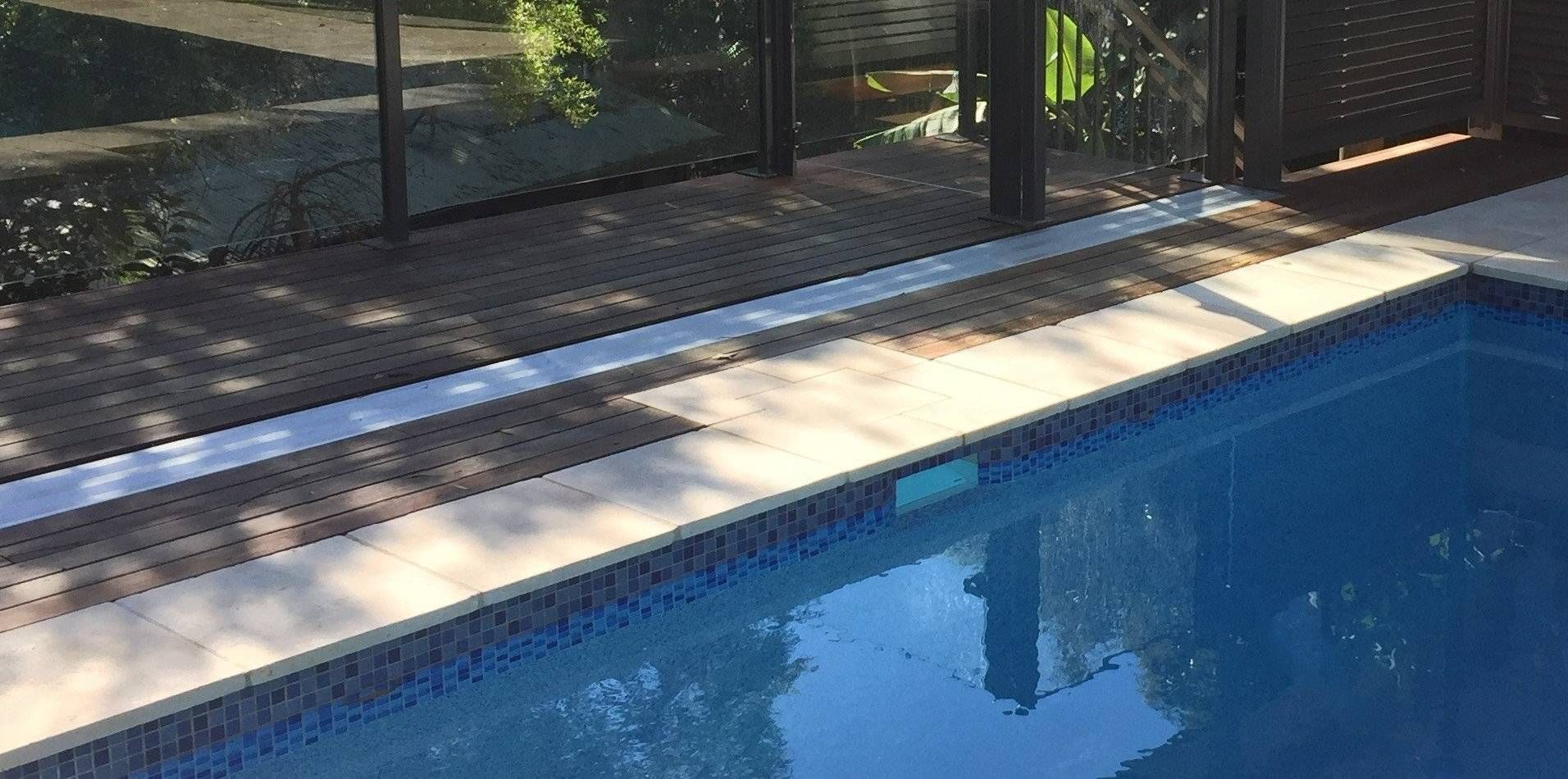 A pool with blue water and tiled edges next to a wooden deck with a fence.