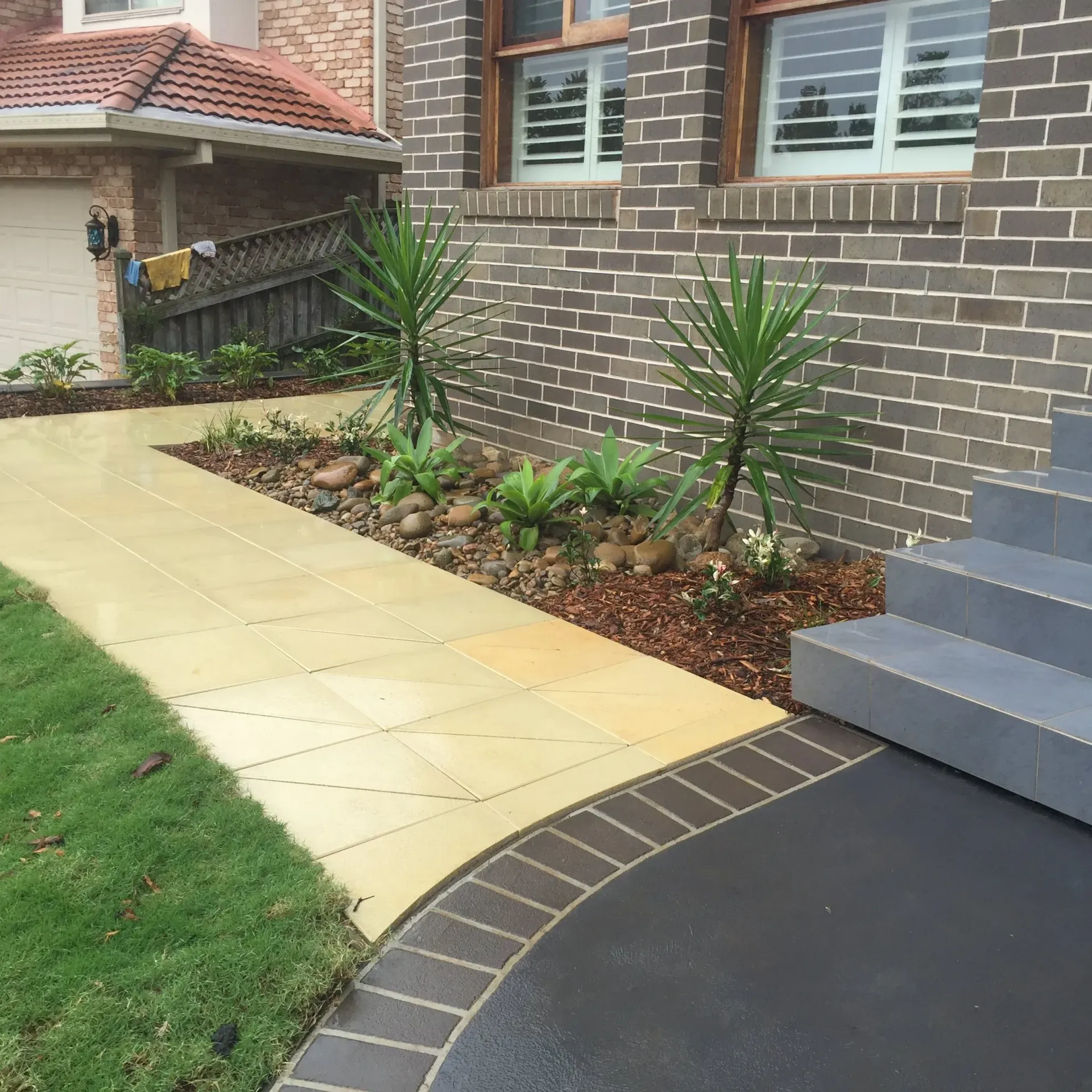 Pathway with landscaping, steps, and house facade. Yellow pavers, dark brick, and greenery.