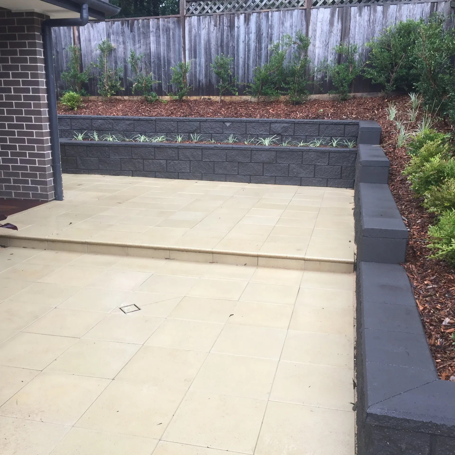 Patio with beige tiled flooring, dark gray retaining walls, and planted greenery. A wooden fence is in the background.
