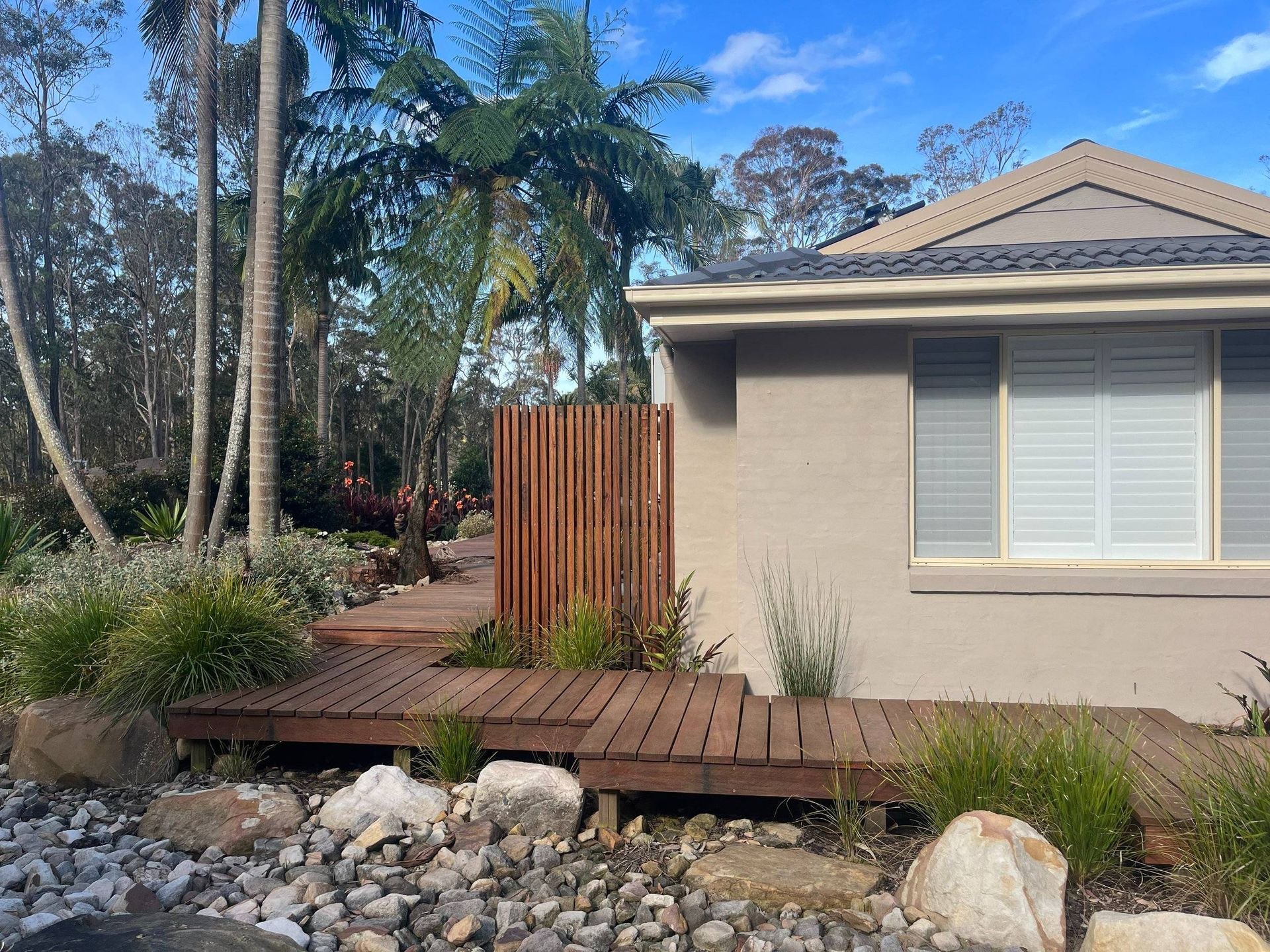 Wooden deck and fence alongside a tan house, surrounded by lush greenery and rocks.