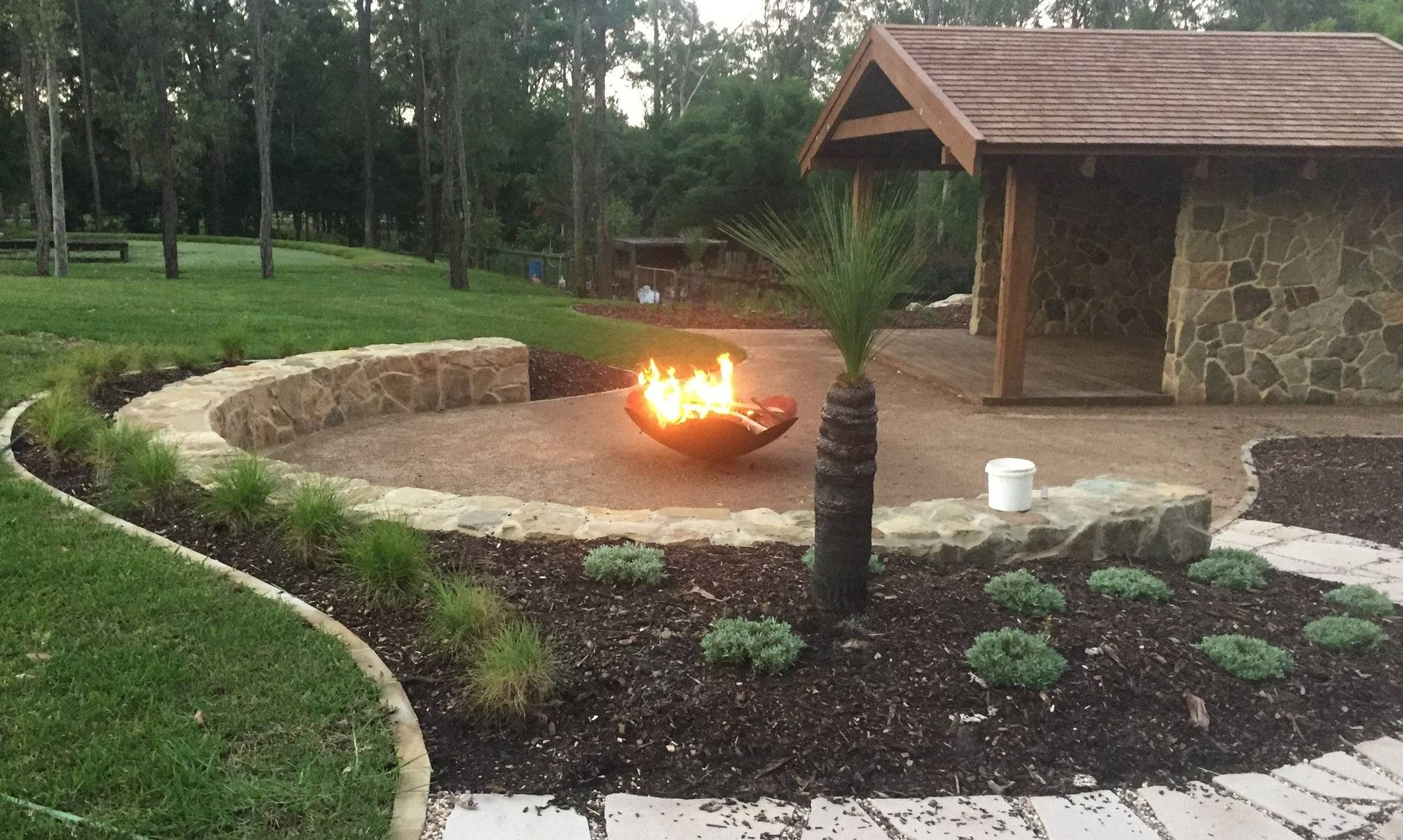 Outdoor fire pit area with stone walls, a small building, and landscaping.