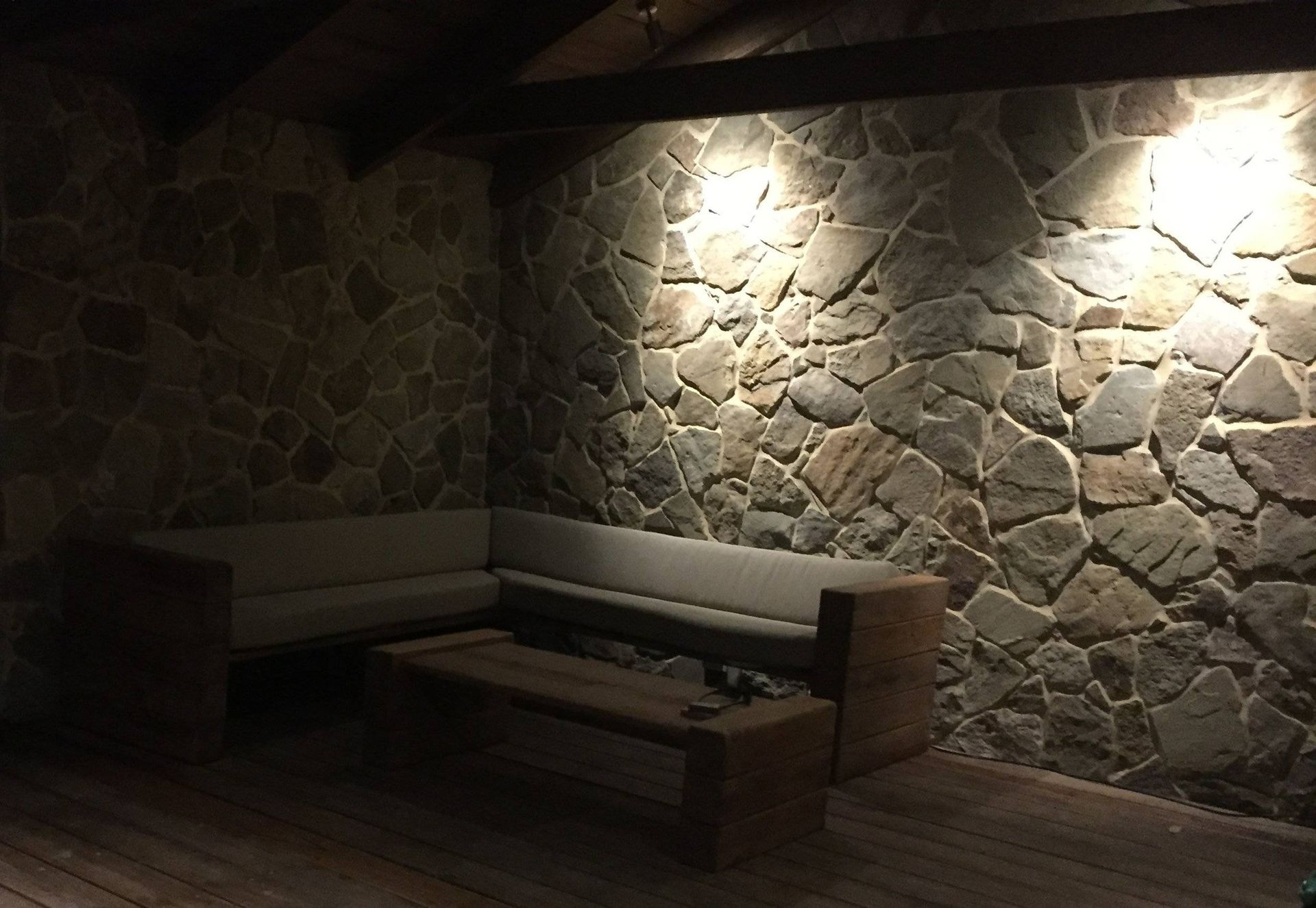 Stone-walled outdoor seating area. Wooden sofa and coffee table are lit by overhead spotlights, creating shadows.