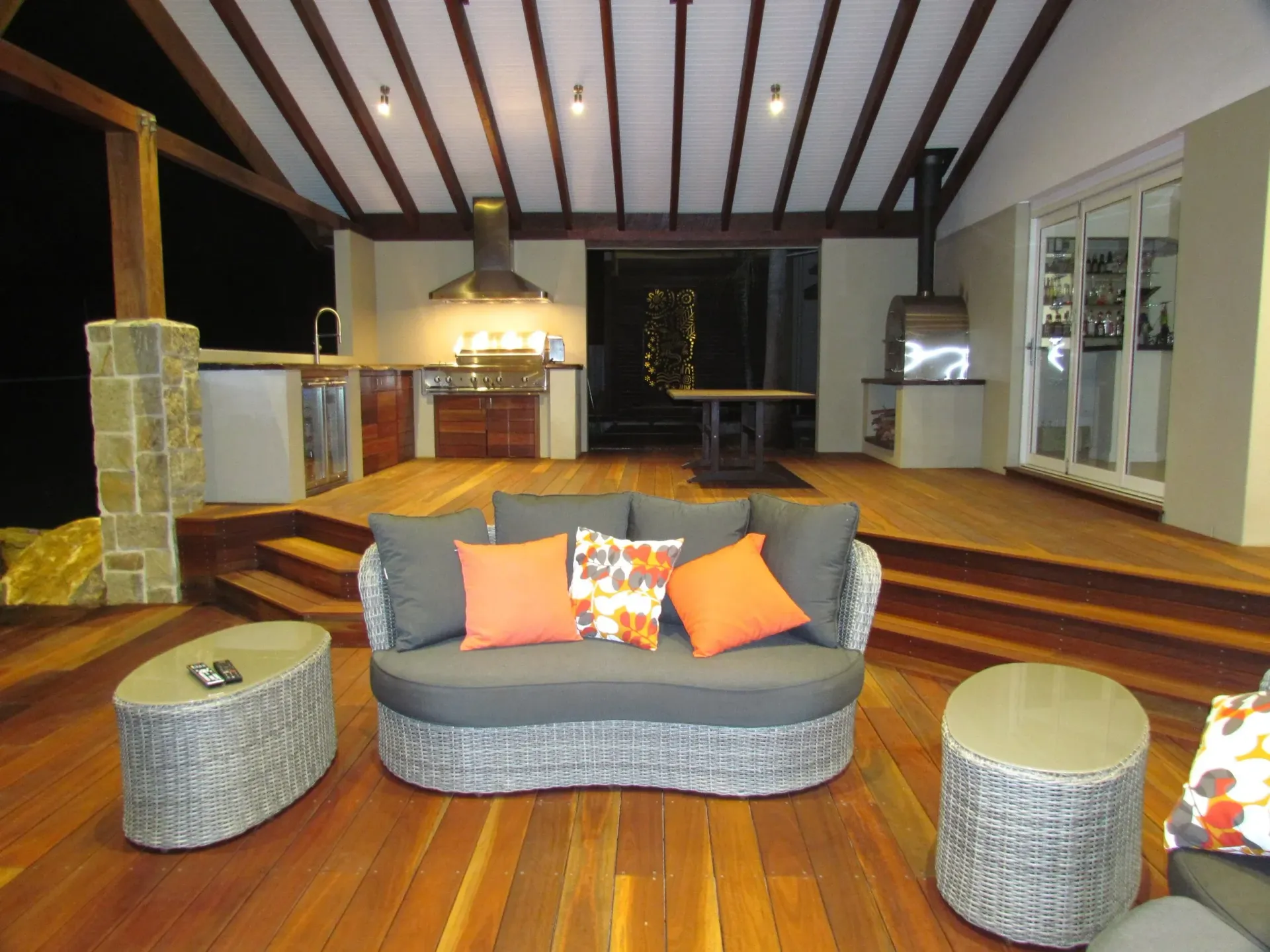 Outdoor living area with sofa, orange pillows, wooden deck, built-in kitchen, and overhead lighting.
