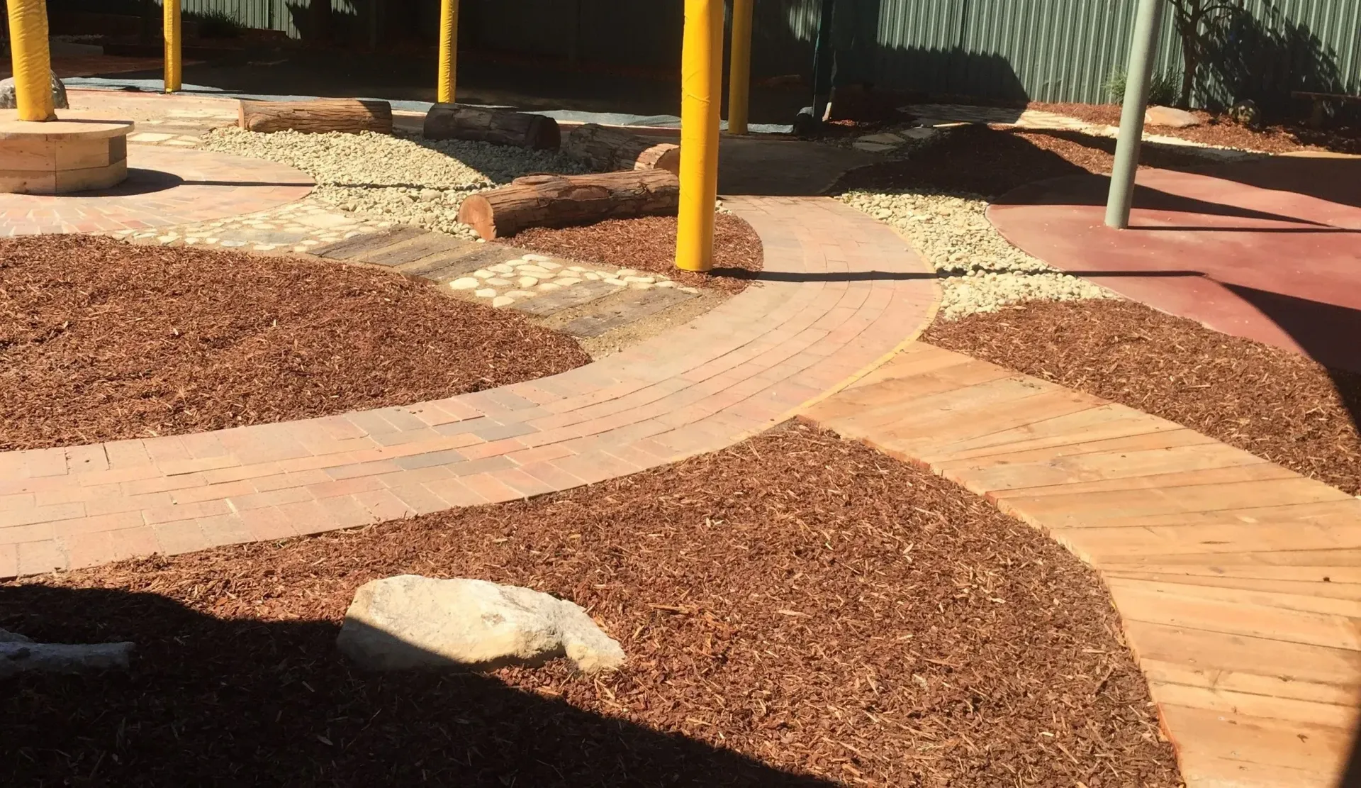 Playground with wood chip groundcover, brick pathways, and yellow support poles.