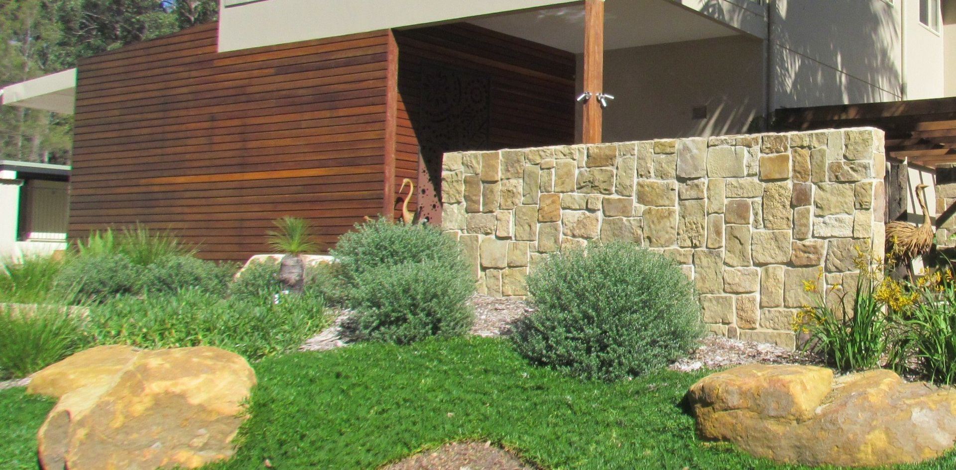 Exterior landscaping with stone wall, greenery, and large boulders in front of a modern building with wooden siding.