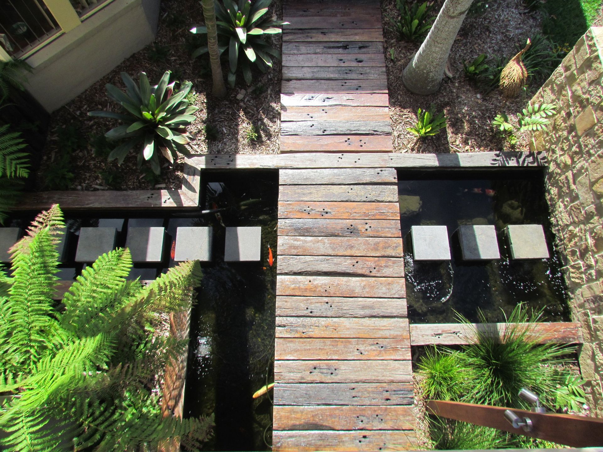 Overhead view of a wooden walkway and stepping stones over a koi pond with surrounding greenery and trees.