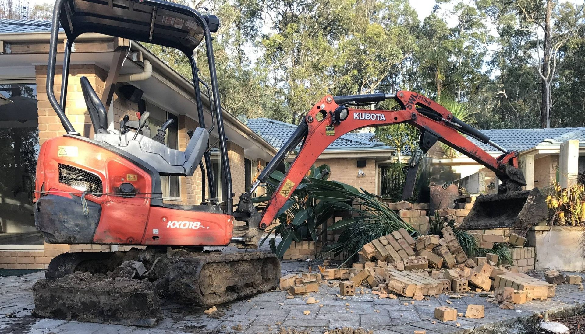 Orange excavator removing bricks next to a house with blue roof.