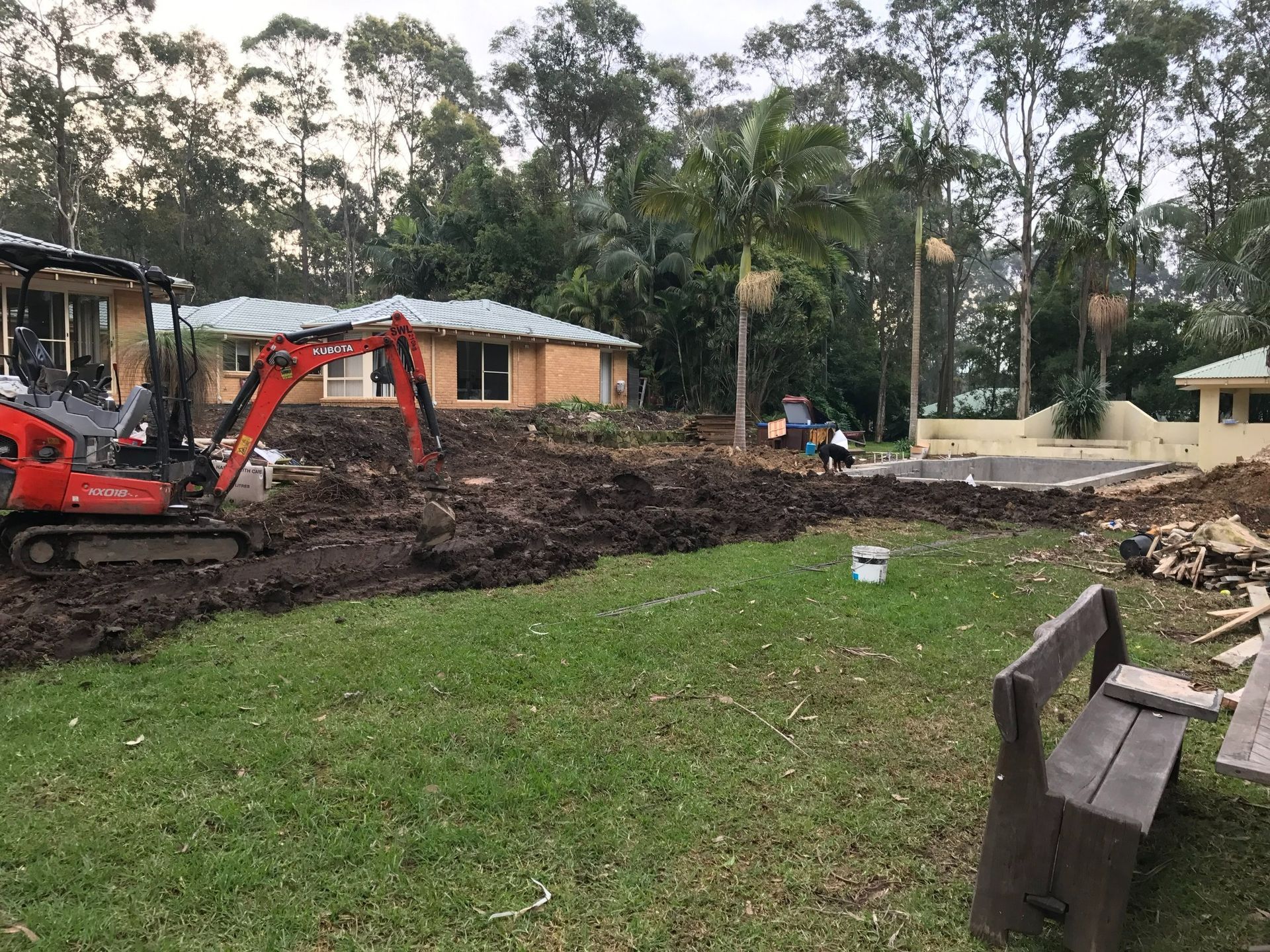 Mini excavator digging in a backyard with houses, trees, and a pool in the background.