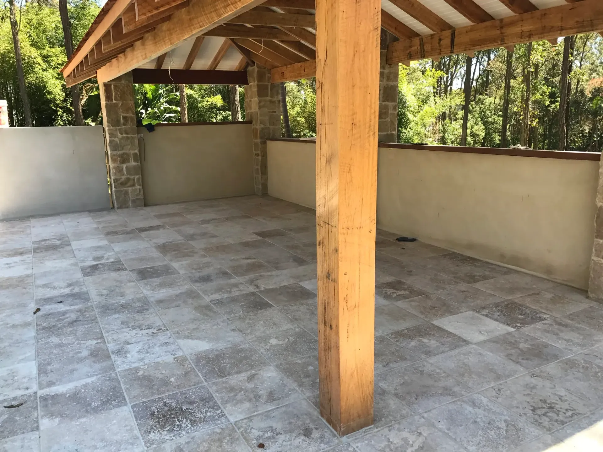 Covered outdoor patio with stone floor and wooden support beams, surrounded by low walls.