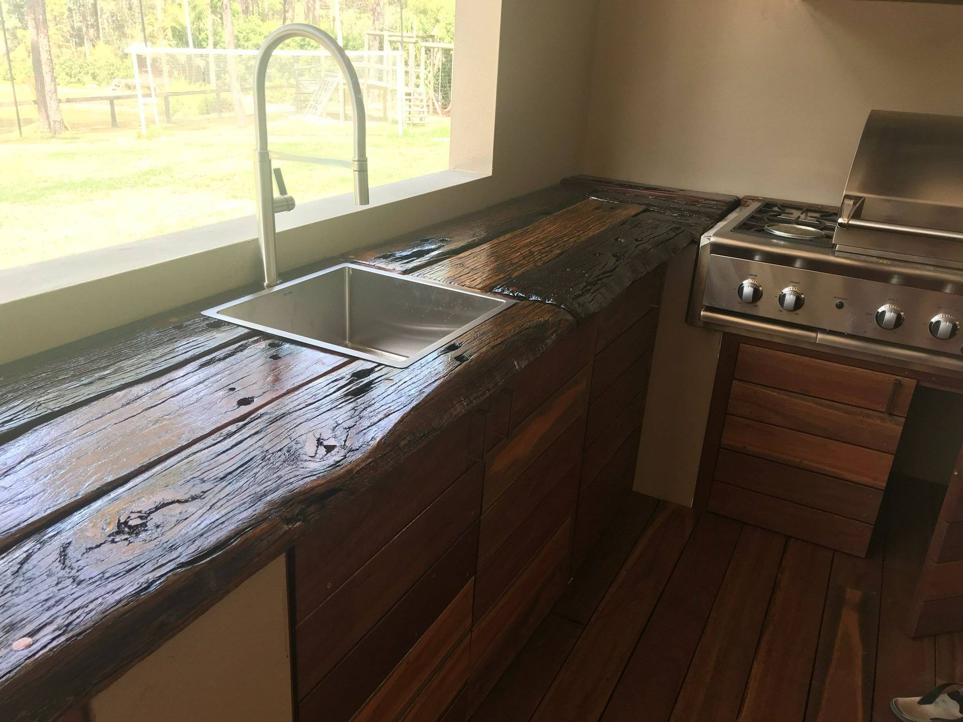 Outdoor kitchen with wooden countertop, stainless steel sink, and built-in grill next to a window.