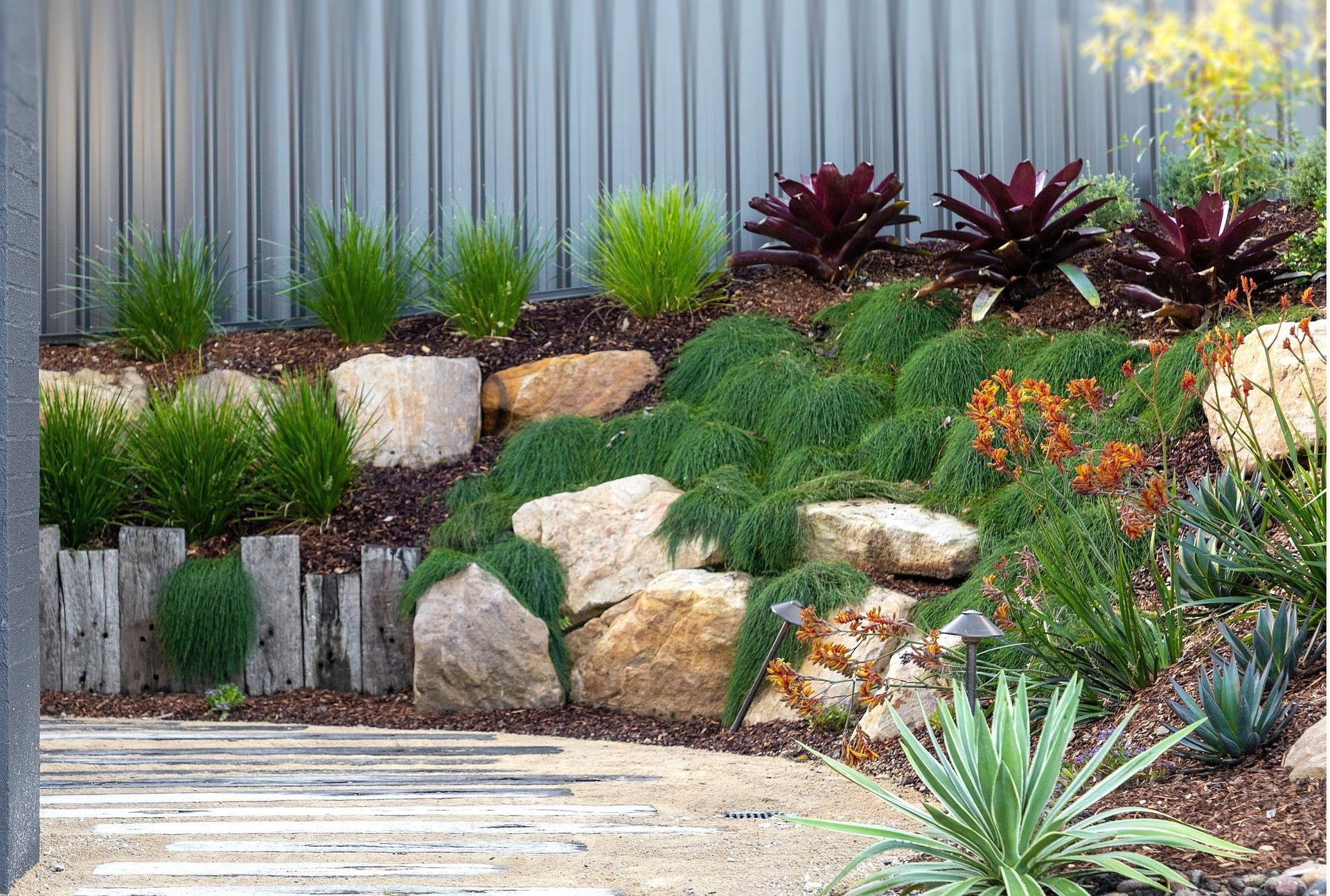 A layered garden bed with rocks, lush green plants, and a corrugated metal fence backdrop.