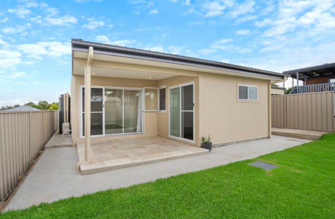 Tan-colored backyard studio with a patio, concrete path, and green lawn. Fenced on both sides, blue sky.
