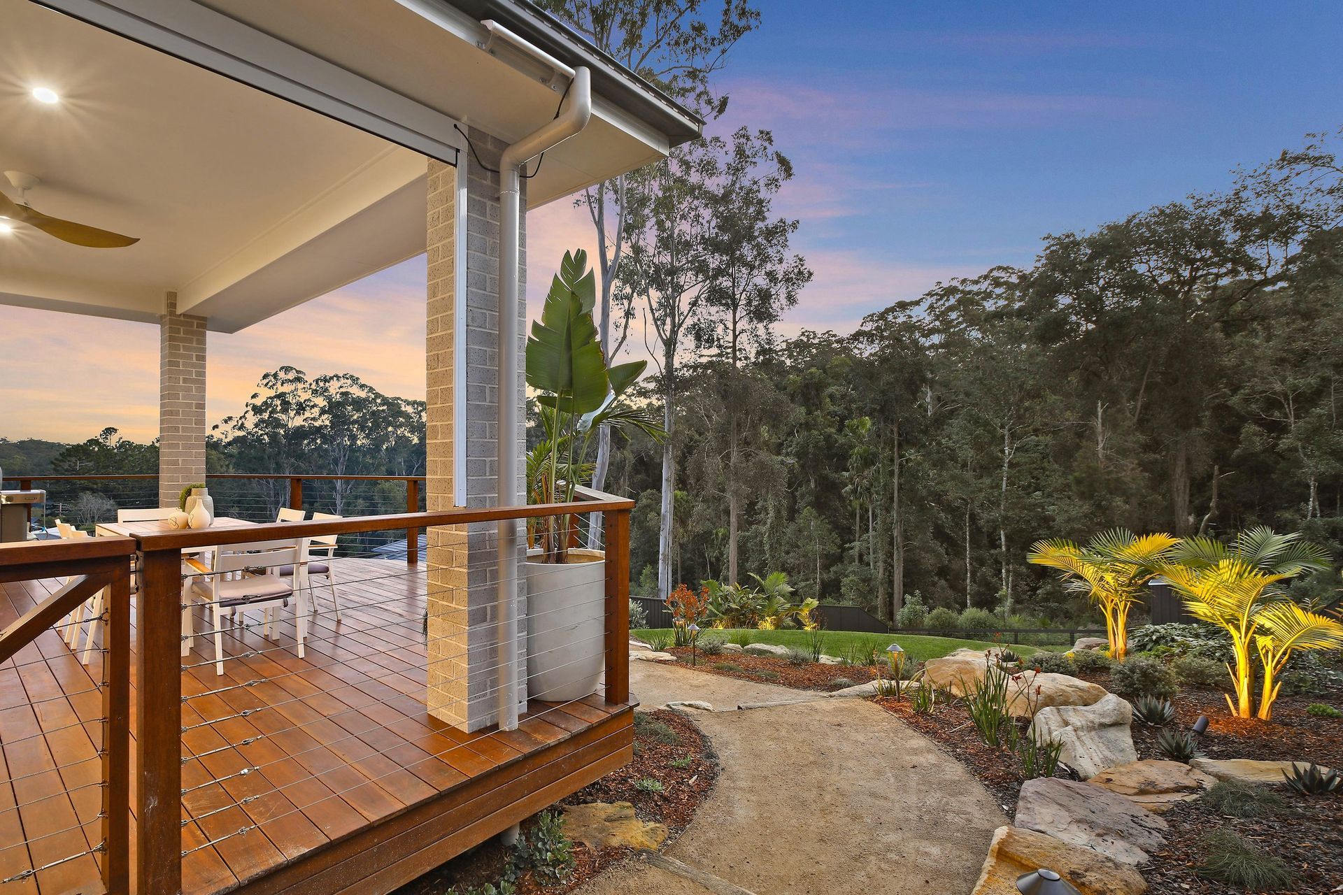 Deck overlooking a lush backyard with a pathway leading to illuminated palm trees, at dusk.