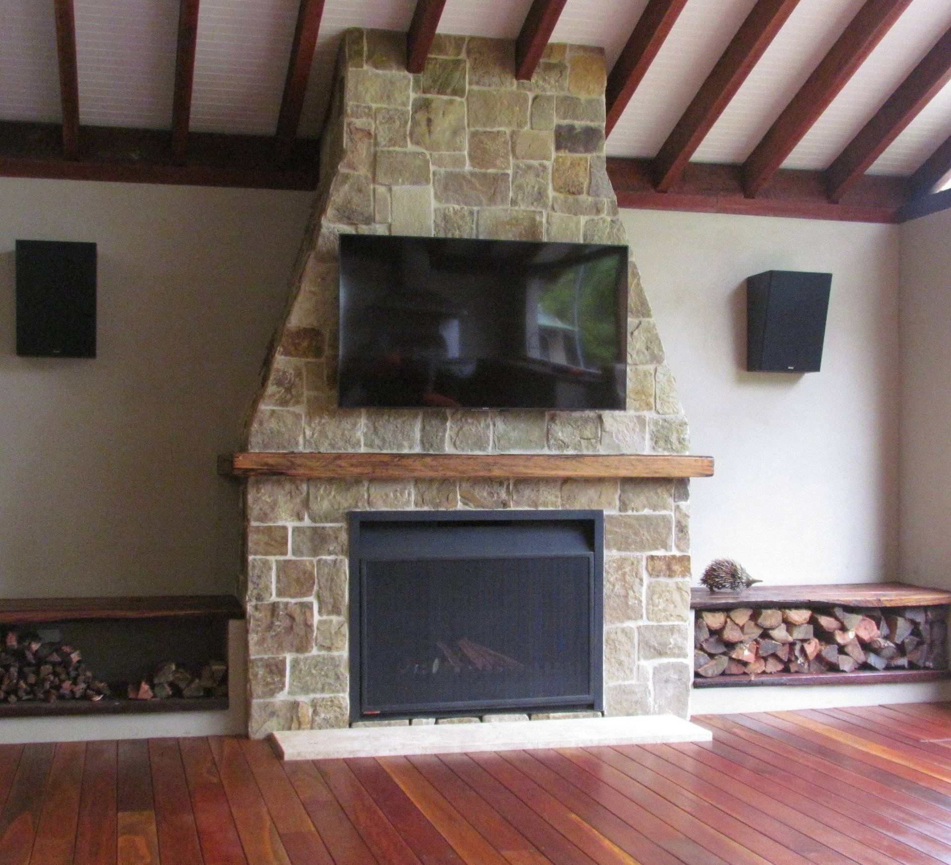 Stone fireplace with a mounted TV, flanked by speakers and wood storage. Wooden ceiling, floor.