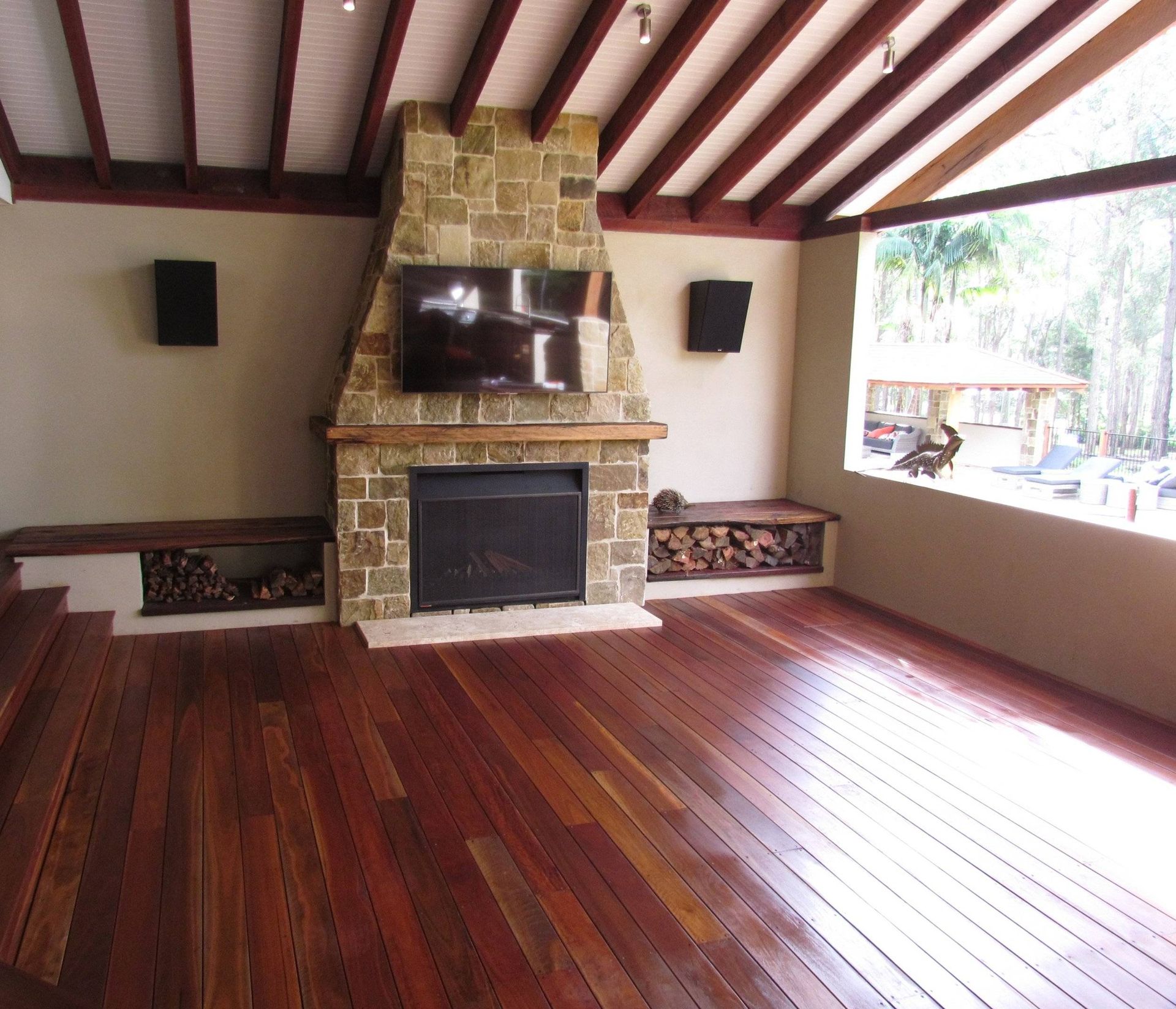 Spacious room with a fireplace, TV, and wood floor. Firewood stored on ledges. Exposed wood ceiling, large window.