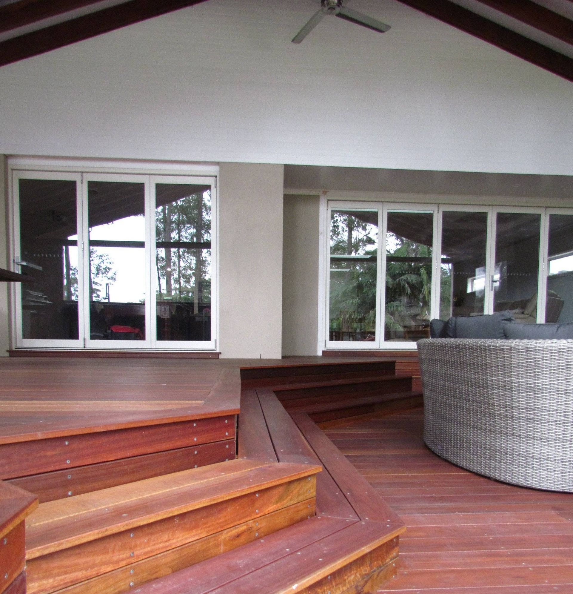 Wooden deck with stairs leading to a covered patio. White framed glass doors and a woven circular sofa are visible.
