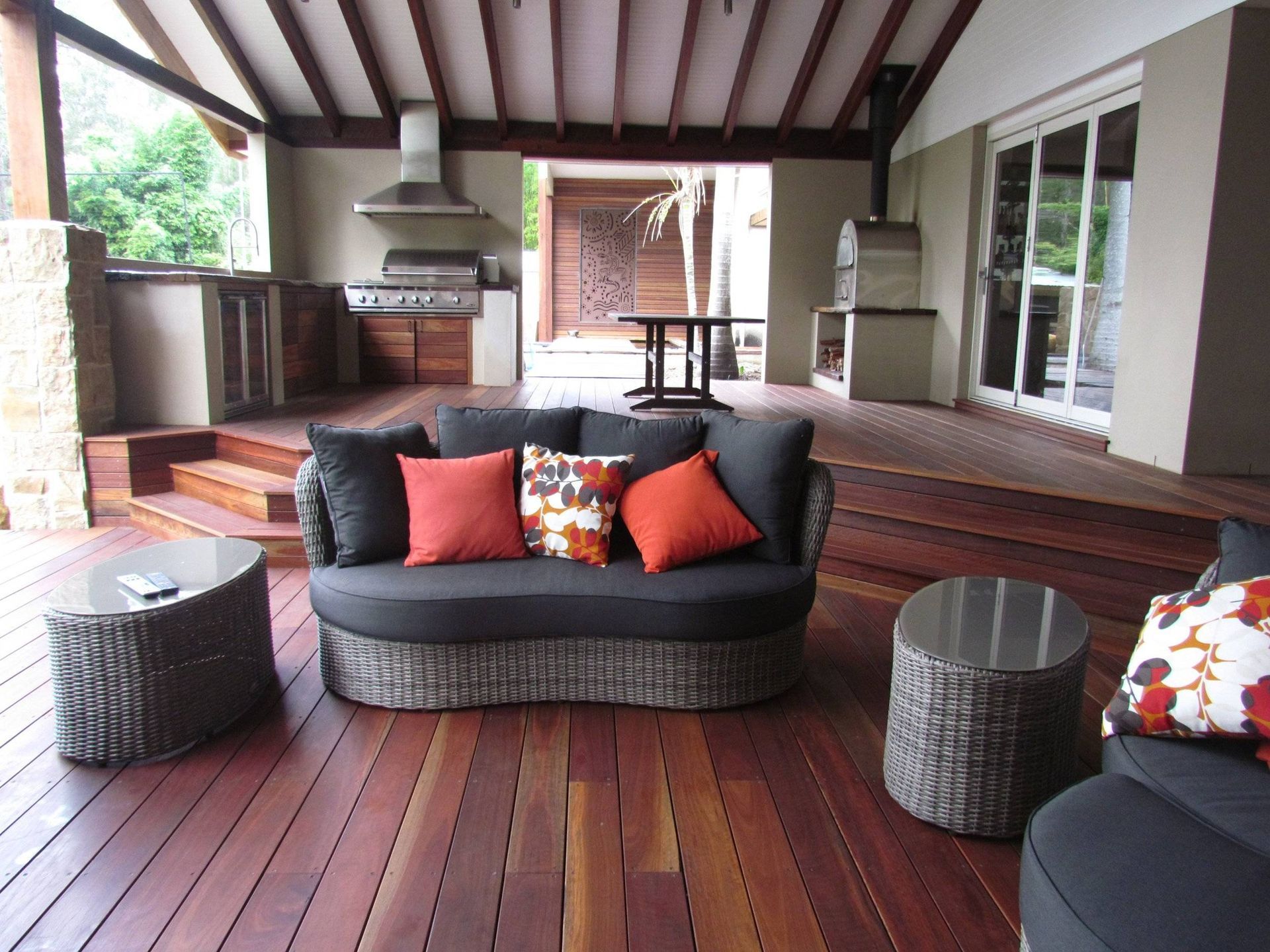 Outdoor patio with dark wood deck, seating area with sofa and colorful pillows, and built-in grill.