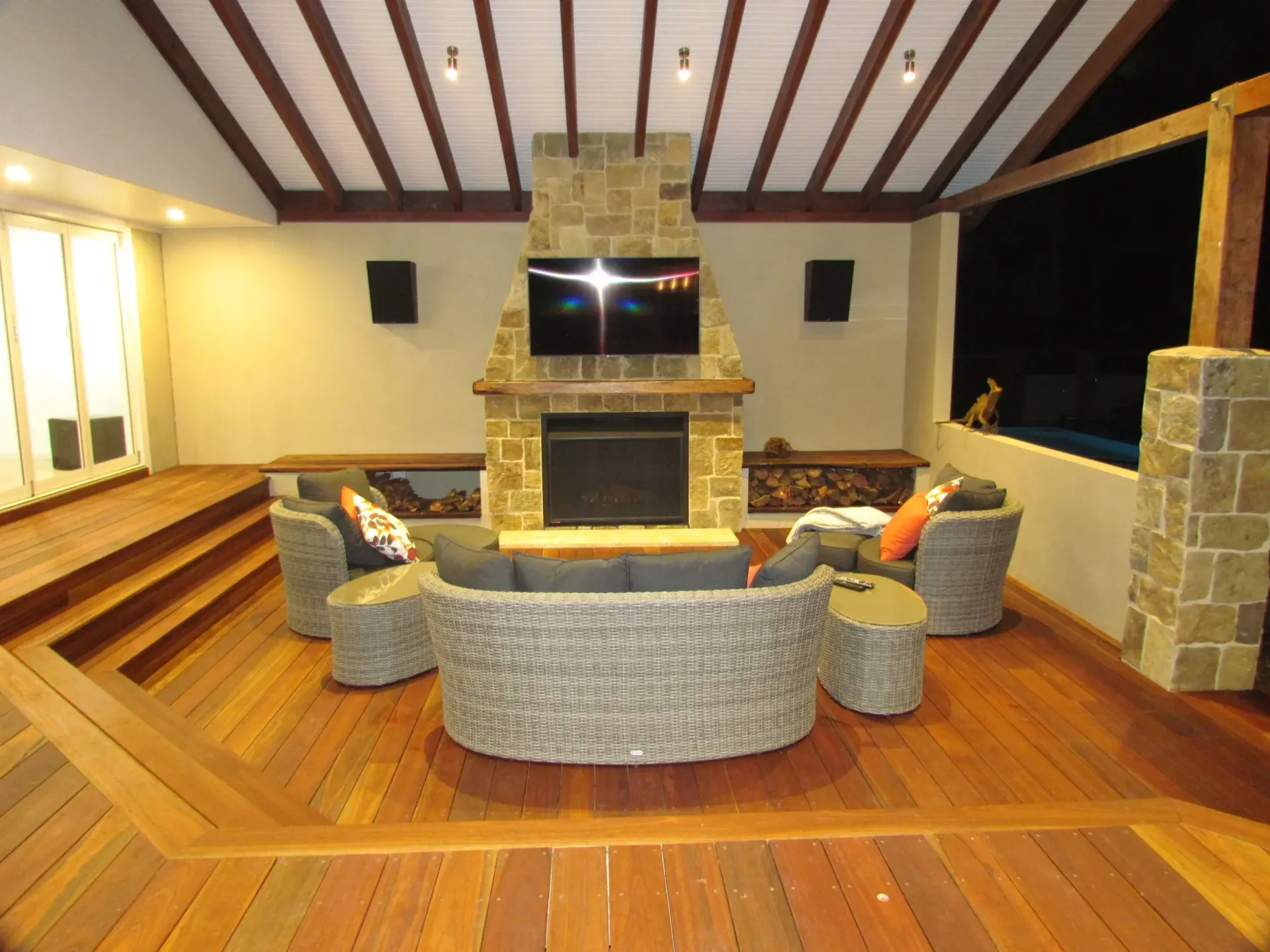 Outdoor seating area with fireplace, TV, and wooden deck. Wicker sofa, armchairs, and wood beams.
