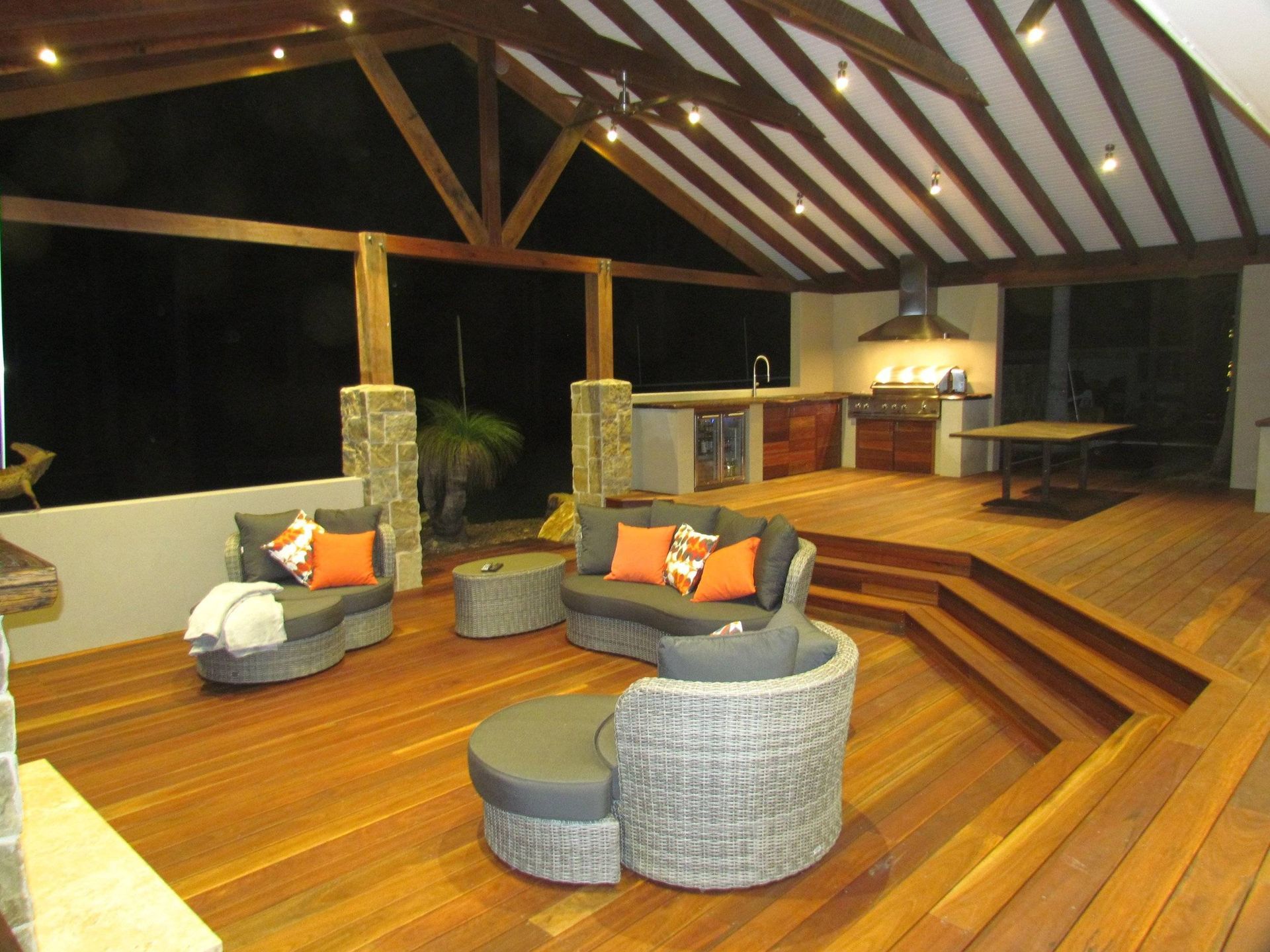 Outdoor kitchen and lounge area with wooden deck, seating, and overhead lighting.